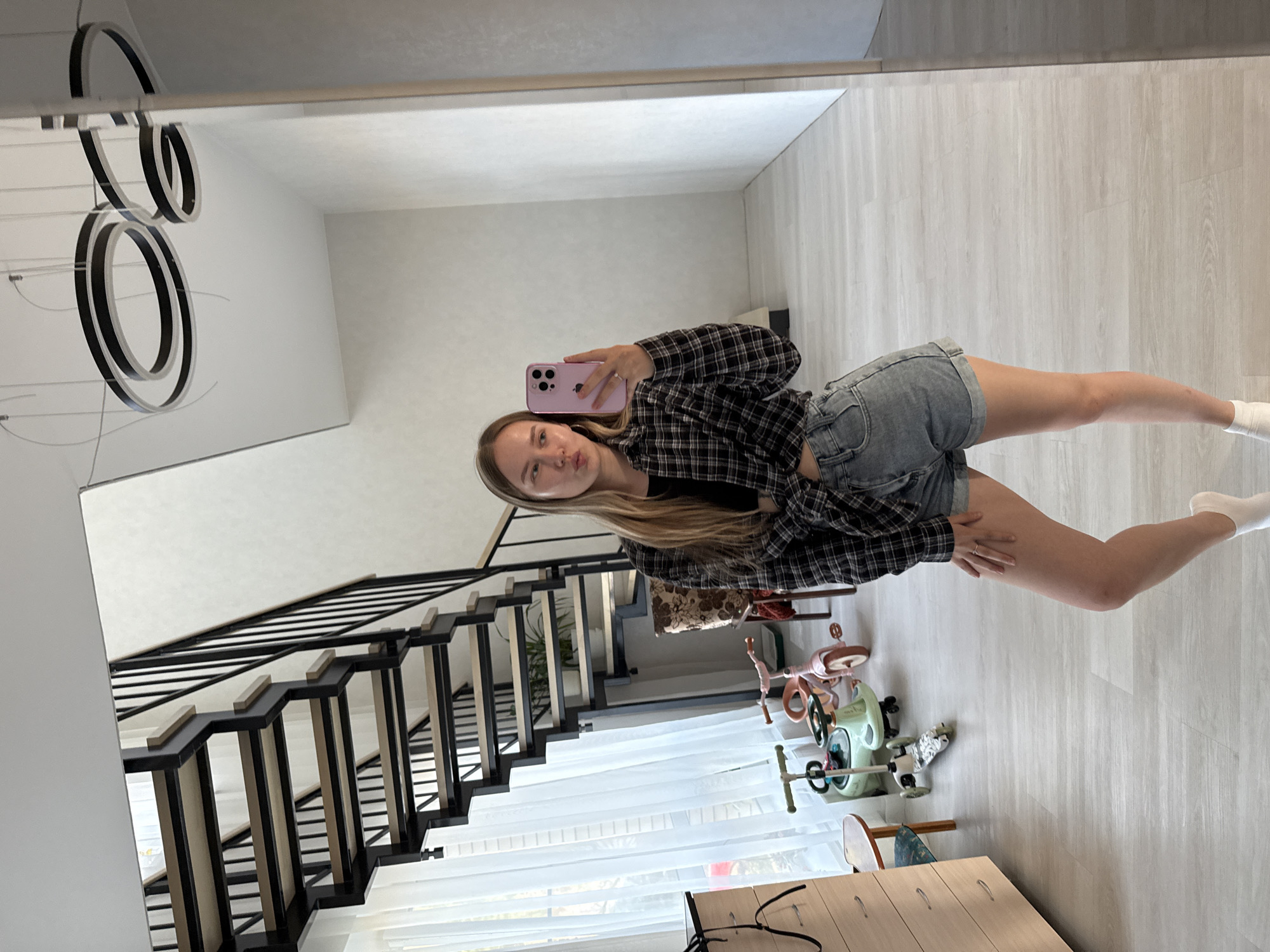 A woman is taking a mirror selfie in what appears to be the hallway of a home. She is wearing a black and white plaid shirt, denim shorts, and white socks. She is holding a pink cell phone. A staircase with black railings is visible in the background, as well as sheer white curtains. There is children’s riding toys in the background. The location cannot be determined from this image alone.