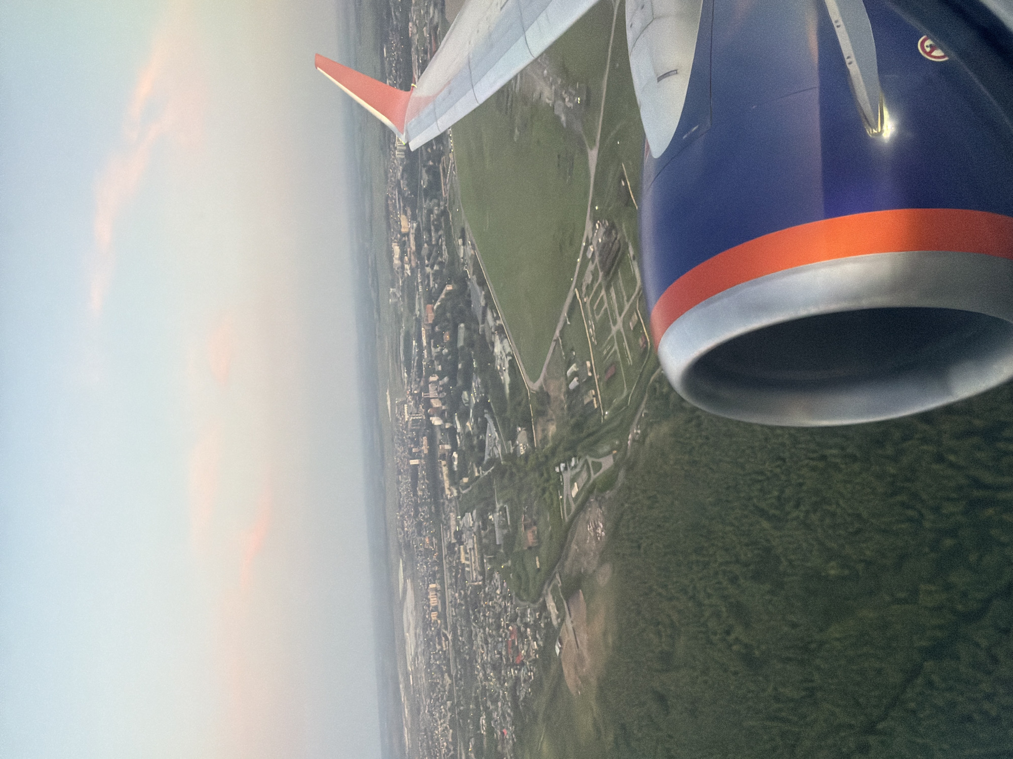 This aerial photograph, taken from an airplane, captures a view of the city of Ob, Russia. In the foreground, the engine of the aircraft is visible, painted in dark blue with orange and silver accents. The wing of the plane, with its distinctive red-tipped stabilizer, extends into the frame on the right. Below, a sprawling urban landscape unfolds. The city features a mix of residential areas with houses and apartment buildings, interspersed with green spaces and industrial zones. A large, empty green field dominates a portion of the mid-ground. Further in the distance, the cityscape extends towards a hazy horizon under a pale blue sky with faint pink clouds, indicative of either dawn or dusk. The overall scene suggests a tranquil descent or ascent over a moderately sized Russian city.