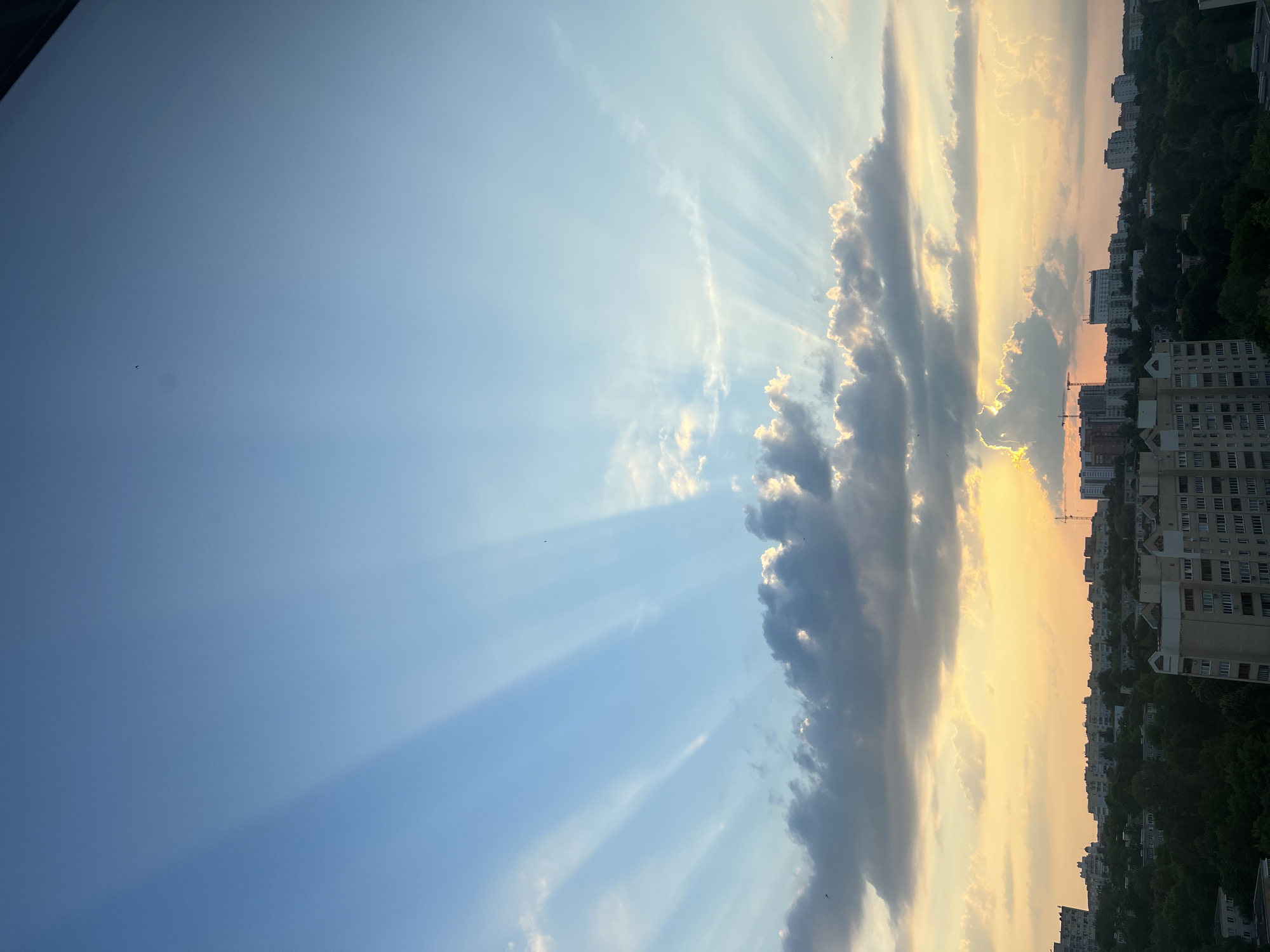 The image depicts a cityscape at sunset, likely in Minsk, Belarus, given the architectural style and urban density. The sky is a prominent feature, dominated by a gradient of blue from the upper half to a warm yellow and orange near the horizon. Dramatic crepuscular rays, shafts of sunlight piercing through the atmosphere, fan out from behind a line of clouds, creating a striking visual effect.
Below the sky, a dense urban landscape unfolds. Numerous multi-story apartment buildings, typical of Soviet-era or post-Soviet construction, are visible, interspersed with areas of lush green trees. The buildings vary in height and architectural detail, with some featuring balconies and visible window patterns. The presence of at least one construction crane suggests ongoing development within the city.
The overall atmosphere is one of a tranquil evening, with the natural beauty of the sunset contrasting with the man-made structures of the city. There are no visible people or animals in the frame, and the focus is on the interplay of light, clouds, and the urban environment. The image is taken from an elevated perspective, possibly from a residential building, offering a panoramic view of the city.