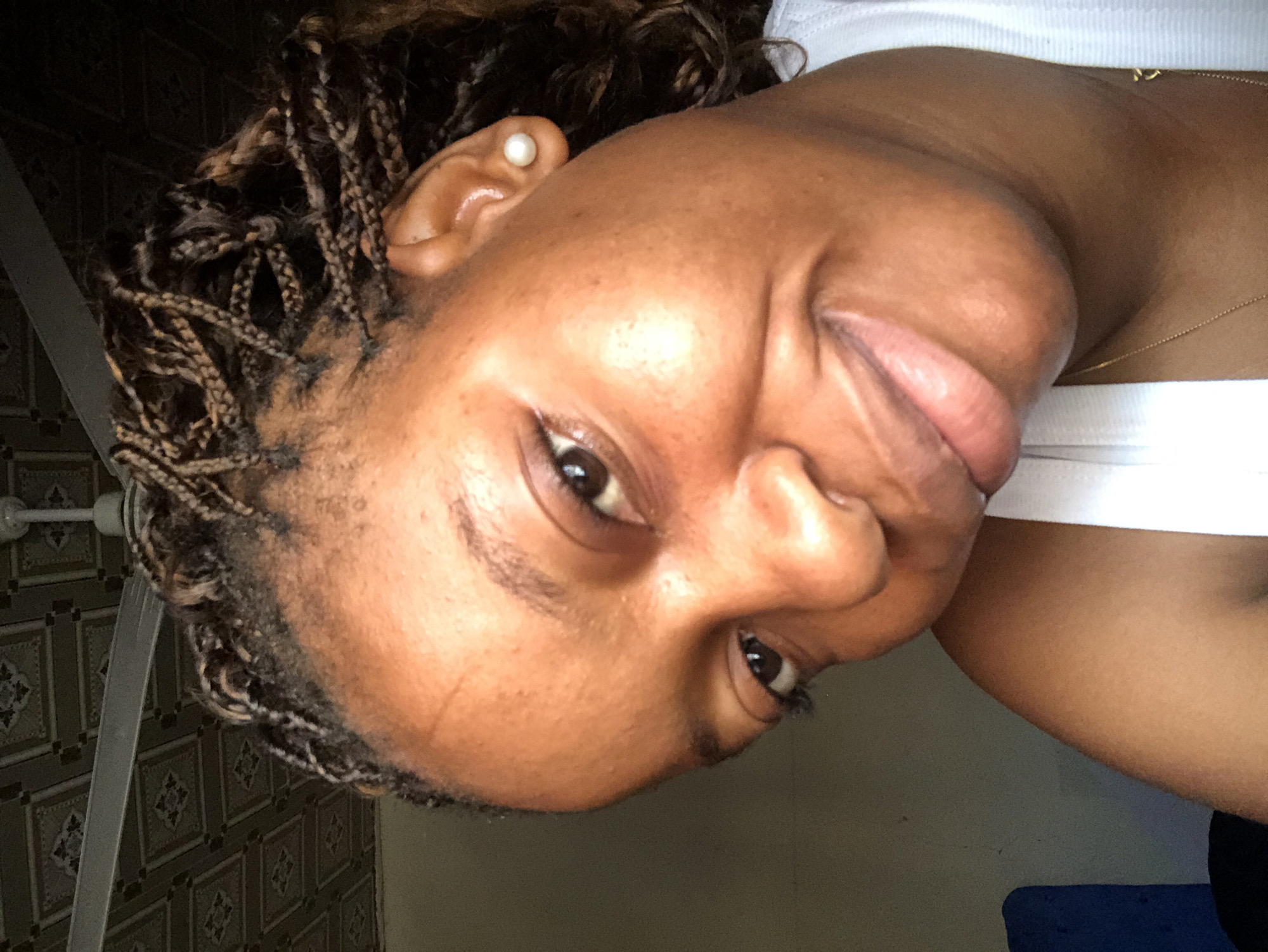 A close-up, rotated portrait of a dark-skinned person with braided hair and a pearl earring in their left ear. The person is wearing a white tank top and a gold necklace. The background shows a patterned wall and a ceiling fan. No definitive information indicates the location is Kashere, Nigeria.