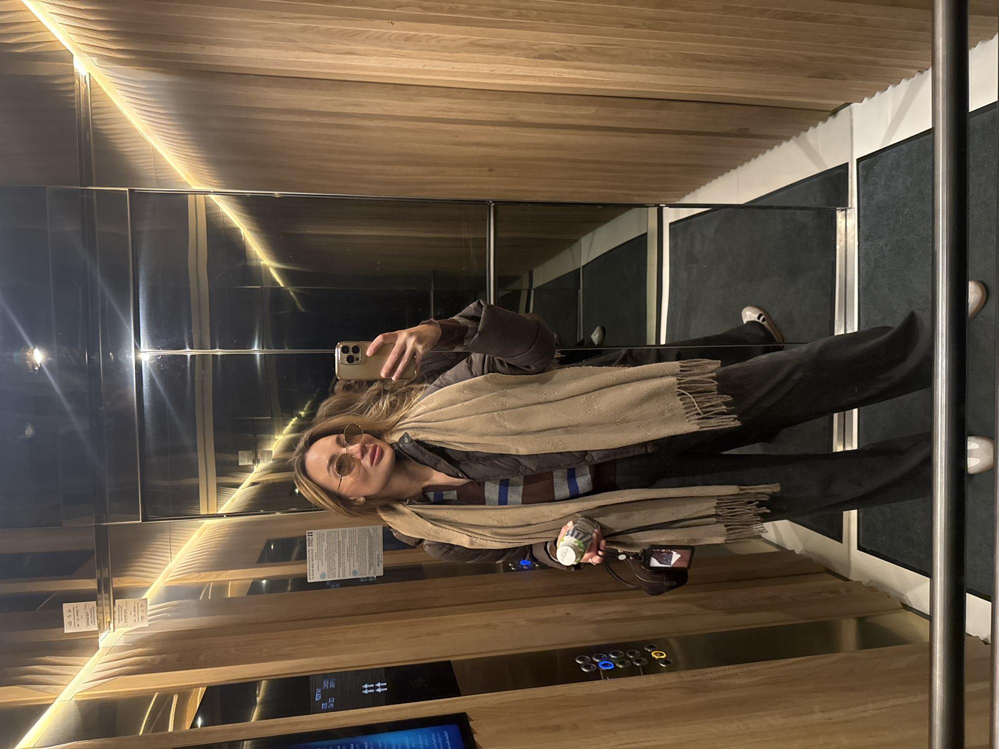 A woman is taking a selfie in an elevator. The elevator has wood-paneled walls and mirrored surfaces. The woman is wearing a scarf and holding a bottle. The elevator has a black floor and a metal handrail. The location in the city is Unknown, Unknown.