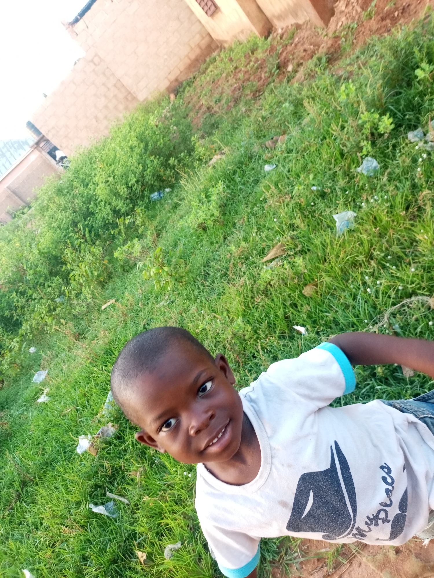 In a high-angle shot taken outdoors, a young Black child is the central focus, positioned in the lower half of the frame. The child is lying down on a patch of green grass, with their head turned towards the camera, smiling. They are wearing a white t-shirt with blue trim on the sleeves and a graphic design on the front that includes text. The background consists of more lush green grass, dirt, and the lower portion of a brick building. The lighting suggests it is daytime, possibly late afternoon due to the shadows. The scene appears to be in a semi-urban or rural area, with hints of a simple dwelling and natural elements. The image has a shallow depth of field, blurring the background to emphasize the child.