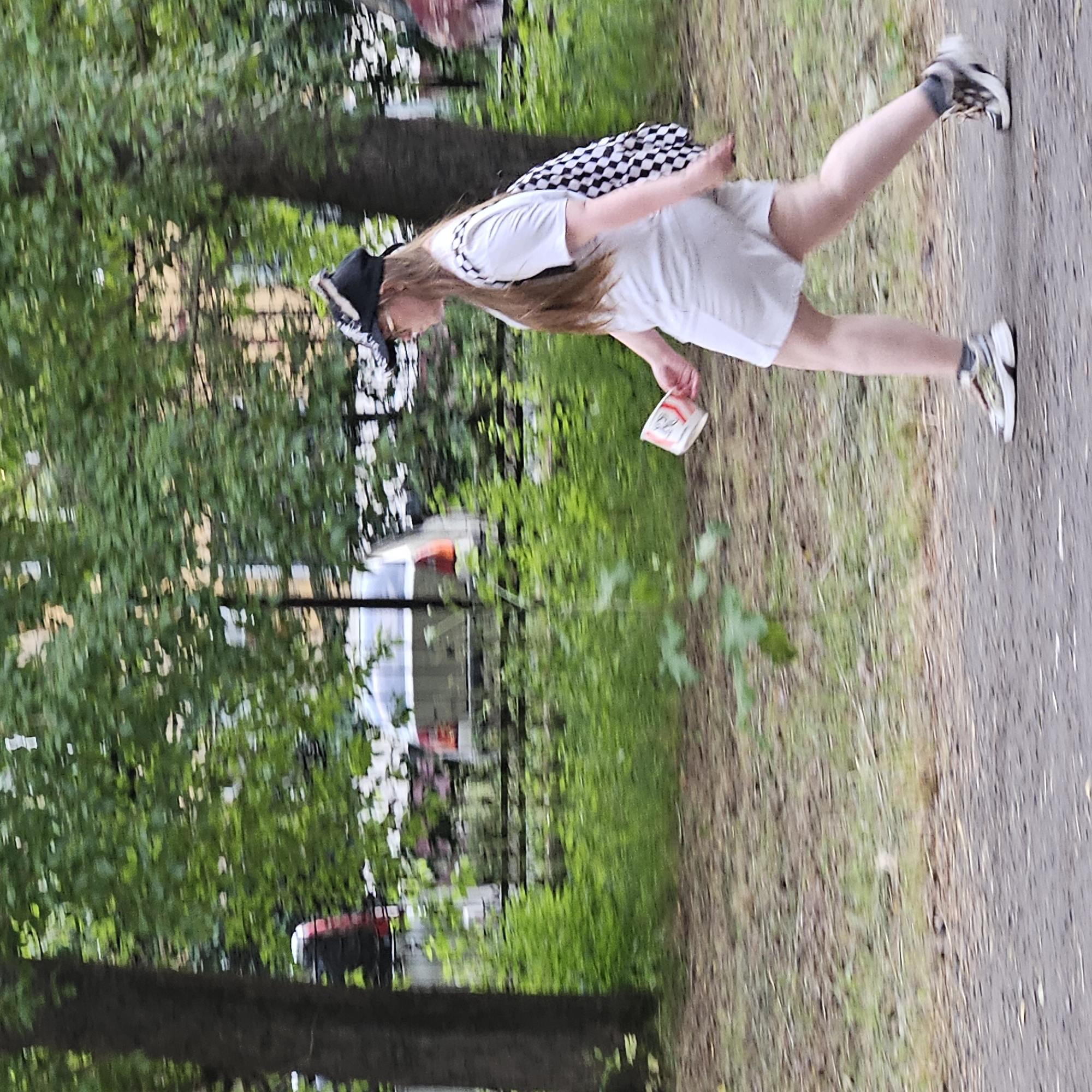 The image shows a young person in motion, likely running or jogging on a paved path. They are wearing a white t-shirt, light-colored shorts, a black and white checkered bandana or headscarf, and sneakers. The person is holding a small, white rectangular object with some red markings in their right hand. The scene appears to be outdoors, with trees and foliage in the background, suggesting a park or a wooded area. The lighting indicates it might be daytime, possibly afternoon, with overcast or dappled light filtering through the leaves. The ground is a mix of dirt, grass, and the paved path. In the background, there are hints of structures, possibly buildings or fences, and some red objects that could be part of a vehicle or play equipment. The image is taken at an angle, and the person is in the mid-ground, captured in a dynamic pose. There is no visible text in the image.