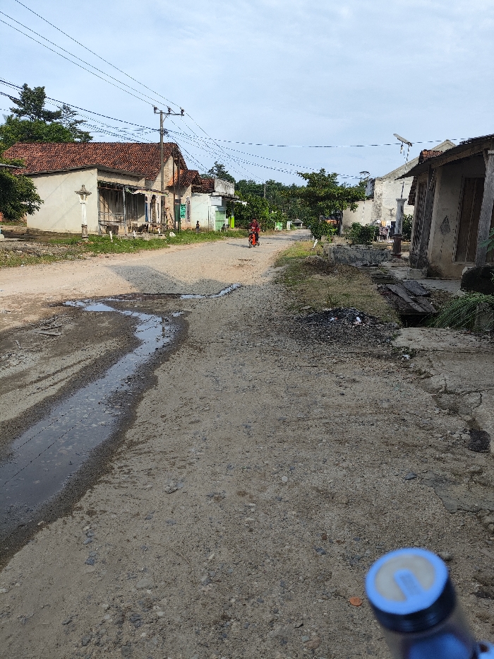 This full shot captures a rural street scene in Kecamatan Selagai Lingga, Indonesia, viewed from a low perspective, likely from a bicycle as evidenced by the blue handlebar end visible in the bottom right corner. The unpaved road, made of dirt and gravel, is central to the image, featuring prominent ruts and muddy puddles, especially along its left edge.
Traditional-style houses with red tiled roofs line both sides of the road. On the left, several weathered houses are visible, with power lines strung across utility poles overhead. A distinctive white, ornate concrete structure, possibly a small monument or grave marker, stands in front of one of these houses. Further down the road, lush green trees and more dwellings are interspersed.
On the right, another set of houses appears, some showing signs of age or disrepair. A satellite dish is mounted on the roof of one of these buildings. The foreground on the right reveals an area with scattered debris, including what appears to be discarded wood planks and other materials.
In the mid-distance, a lone individual, dressed in red clothing, is moving along the road, possibly walking or riding a bicycle or small scooter, adding a human element to the otherwise quiet landscape. The sky is bright and partly cloudy, suggesting it is daytime, perhaps early morning or late afternoon, given the soft, diffused light and the absence of strong shadows. The overall scene depicts a typical, serene moment in a rural Indonesian village. No discernible text is visible within the image.