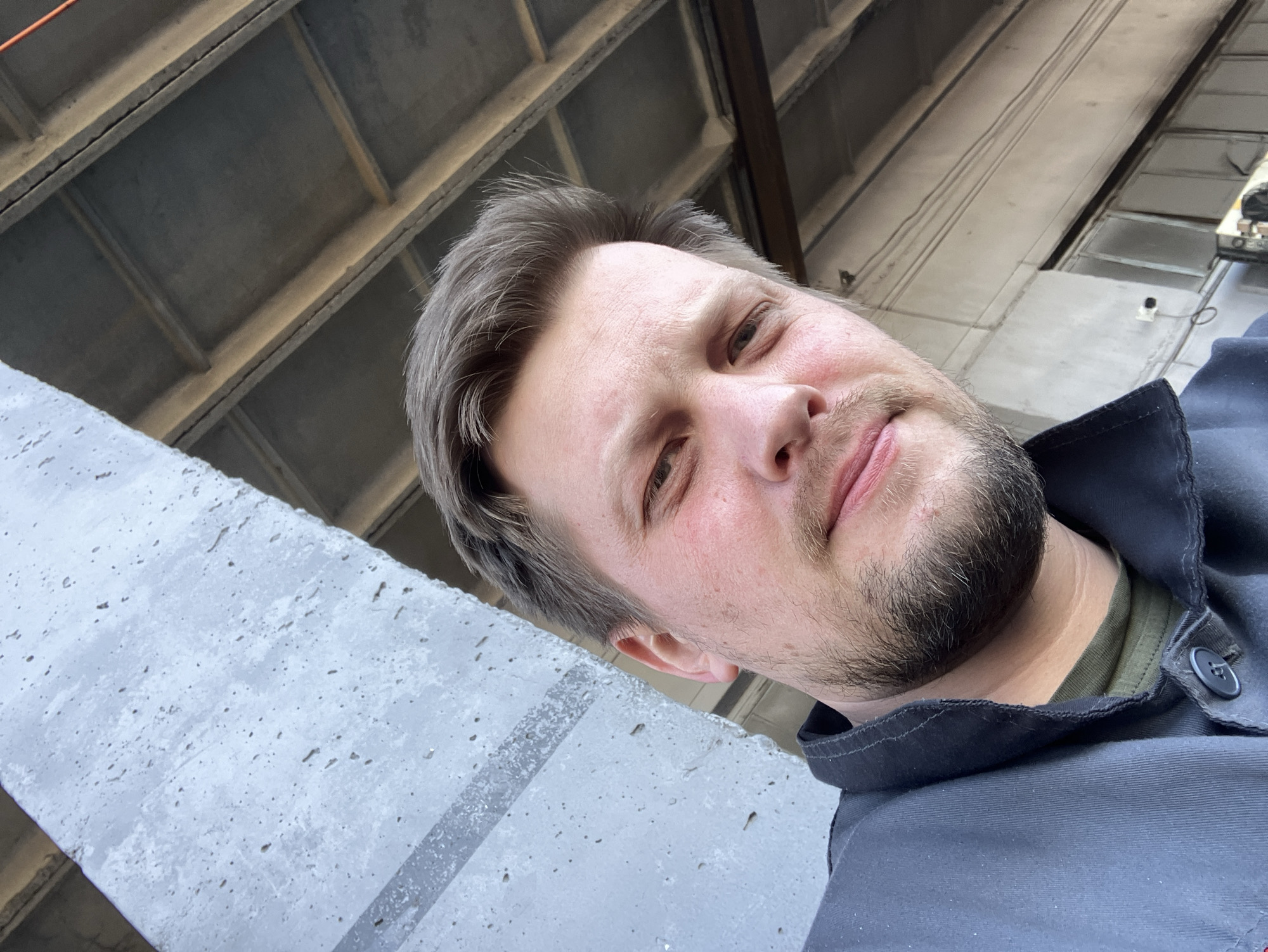 This image presents a close-up portrait of an adult male within an industrial or utilitarian setting in Noviy, Russia. The man, positioned slightly off-center, has light skin, dark blonde hair, and a short beard and mustache. He is wearing a dark grey collared utility jacket over an olive green t-shirt and looks directly at the camera with a neutral expression. To the left, a textured, light grey concrete wall with a horizontal dark grey stripe is visible. The upper right background shows an industrial-style ceiling with corrugated metal panels and exposed support beams. Below the ceiling, to the right of the man, a section of an industrial building wall with vertical paneling is visible, along with a white wall-mounted electrical component and a suspended black wire. A piece of metallic machinery is partially visible in the lower right corner of the frame.
