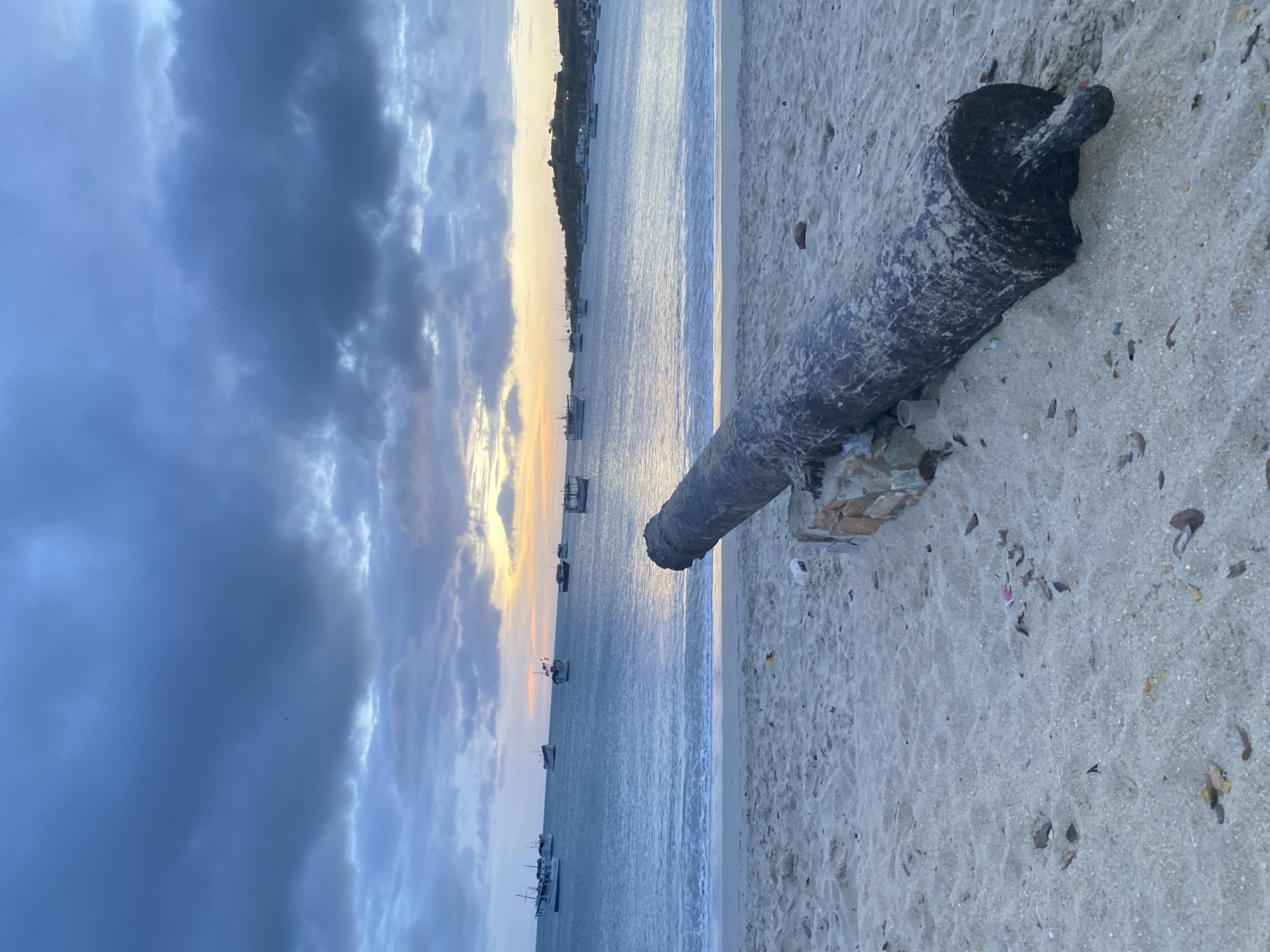 The image depicts a beach scene, potentially at sunset or sunrise, in Juan Griego, Venezuela. The sky is filled with dark, layered clouds in the upper left quadrant, transitioning to a lighter, yellow-tinged sky near the horizon. The water is visible in the middle, with several boats anchored in the distance. A cannon barrel rests on the sandy beach. The barrel is dark and weathered, resting on a base of rocks or bricks. The beach itself is light-colored sand, with some visible debris. The overall scene suggests a coastal environment with remnants of historical artifacts.