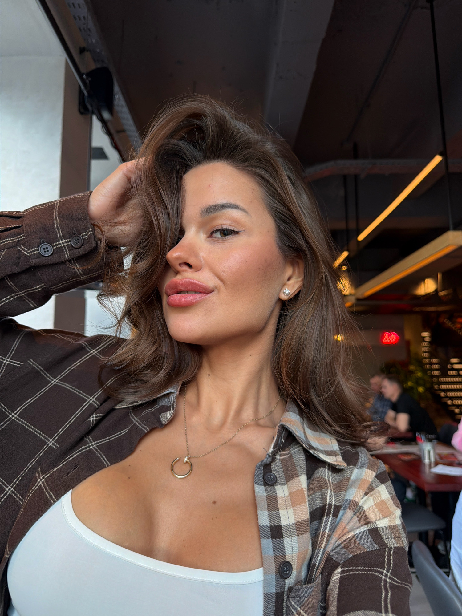 The image is a close-up, selfie-style photograph of a young woman with brown hair and full lips, wearing a white tank top and an open, brown plaid shirt. She is wearing a simple gold necklace with a circular pendant and a small diamond stud earring in her visible ear. Her left hand is raised and touching her hair near her temple. The background suggests an indoor setting, possibly a restaurant or cafe, with industrial-style lighting and other patrons blurred in the distance. The overall lighting is warm, and the focus is on the woman's face and upper body. There is no specific indication of the city of Krasnoyarsk, Russia, within the image itself.