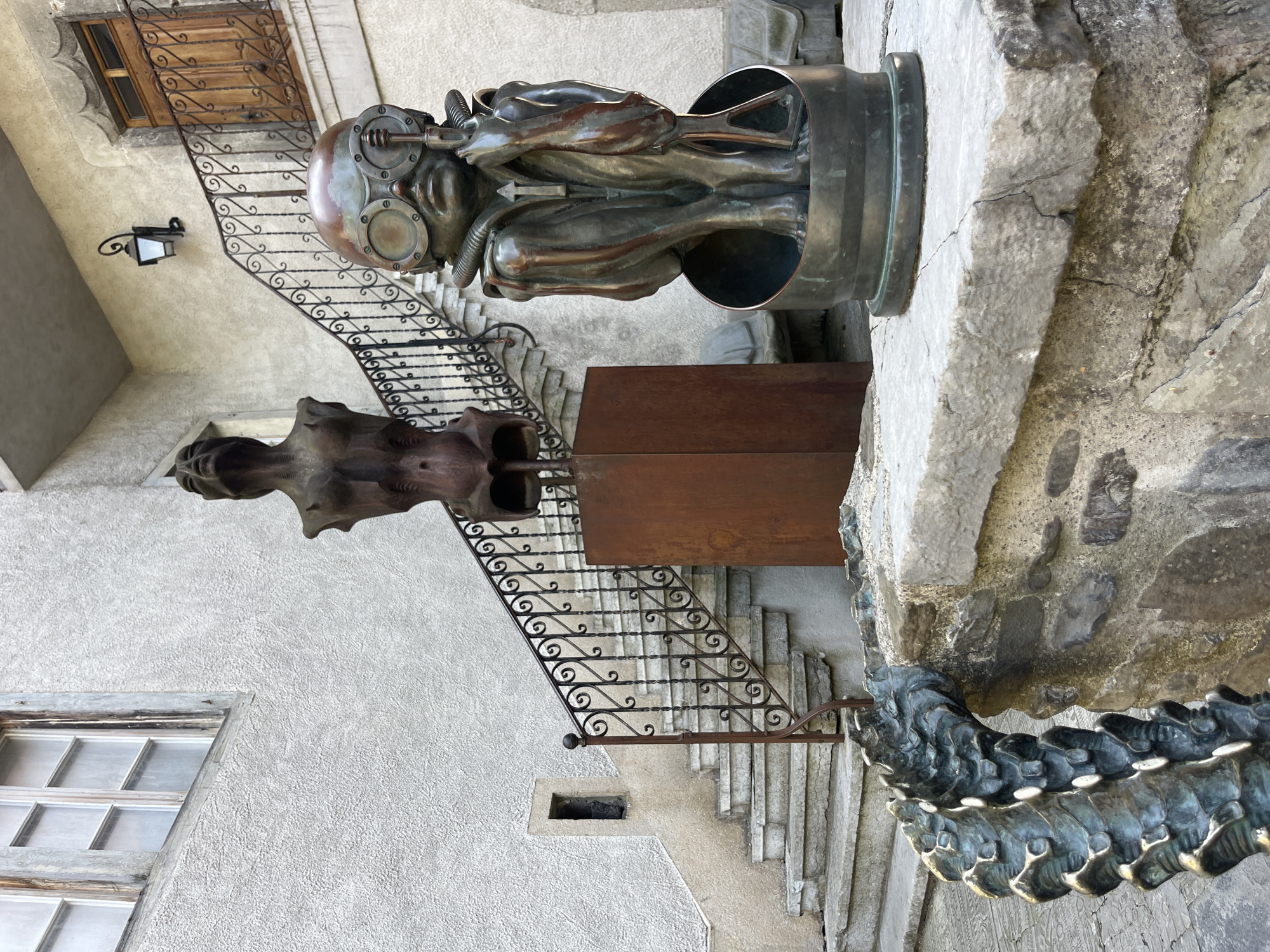The image depicts an outdoor scene with several sculptures and architectural elements. In the foreground, a bronze sculpture of a seated figure wearing goggles and a breathing apparatus is visible. To the left, a portion of another sculpture, resembling a segmented, metallic tail or spine, curves over a stone wall. Behind these sculptures, a stone staircase ascends to a building with a white, textured facade. A wrought-iron railing with ornate scrollwork lines the staircase. Further up the stairs, a wooden door with glass panes and decorative ironwork is set into the building. Above the door, a black, wrought-iron lantern is mounted on the wall. Another sculpture, a bronze torso with pronounced ribs, is displayed on a tall, rusted metal pedestal to the left of the staircase. A window with multiple panes and a weathered wooden frame is visible on the upper left portion of the building. The overall environment suggests a courtyard or outdoor exhibition space within a historic European setting. The city of Epagny, Switzerland, is mentioned as the location, implying the sculptures and architecture are situated there.