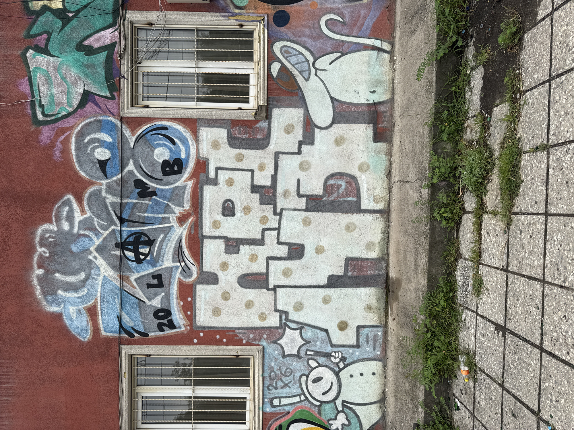 An exterior shot captures a section of a building's red-painted stucco wall adorned with graffiti. Two barred windows with white frames are visible on the left and right sides of the image. The graffiti features stylized lettering and cartoonish figures. On the left, a snowman-like character with a star wand stands next to a set of numbers that read "20 6". In the center, large, white block letters with beige polka dots are prominent, appearing to spell "HERE". To the right of the central lettering, a long-snouted, white cartoon mouse wearing a hat is depicted. The ground in front of the wall is paved with concrete tiles and overgrown with grass and weeds, with scattered debris including cigarette butts and a plastic bottle. The overall scene suggests an urban environment with evidence of neglect. The graffiti style and imagery are common in urban art and street culture. The image was taken in Tbilisi, Georgia.