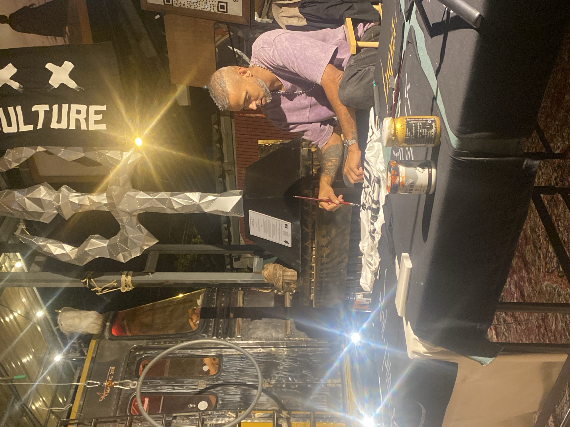 The photograph shows a man with graying hair and a lilac shirt painting on a table covered with a black cloth. He is wearing a necklace and has tattoos on his left arm. Two paint jars are on the table next to him. Above the table hangs a banner that reads "CULTURE" with two X symbols above it. The banner is partially illuminated by a bright light source. An abstract metallic sculpture hangs above the banner. In the background, there is a decorated bus with various attachments and modifications, including a steering wheel and what appears to be a tail light. The image was taken in Thong Sala, Thailand.