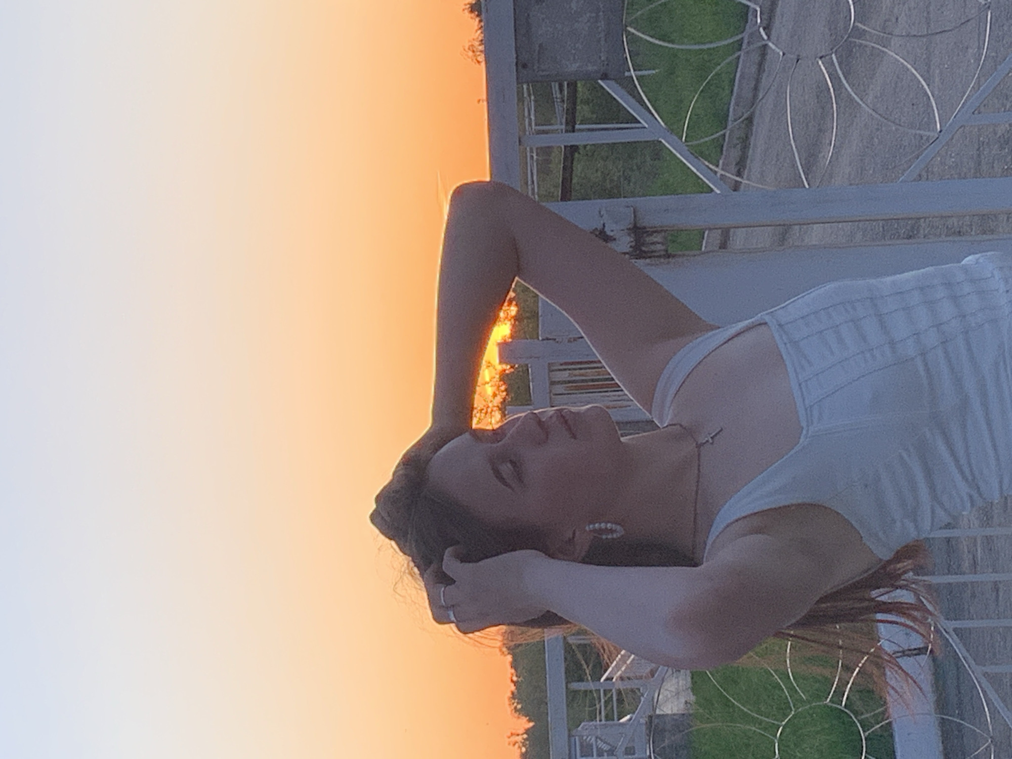 A woman with brown hair is lying on a white metal structure at sunset. The sky transitions from orange to pale blue. The metal structure appears to be part of a decorative fence or railing. Green grass is visible in the background.
I cannot confirm the location within the city of Фоки, Russia.