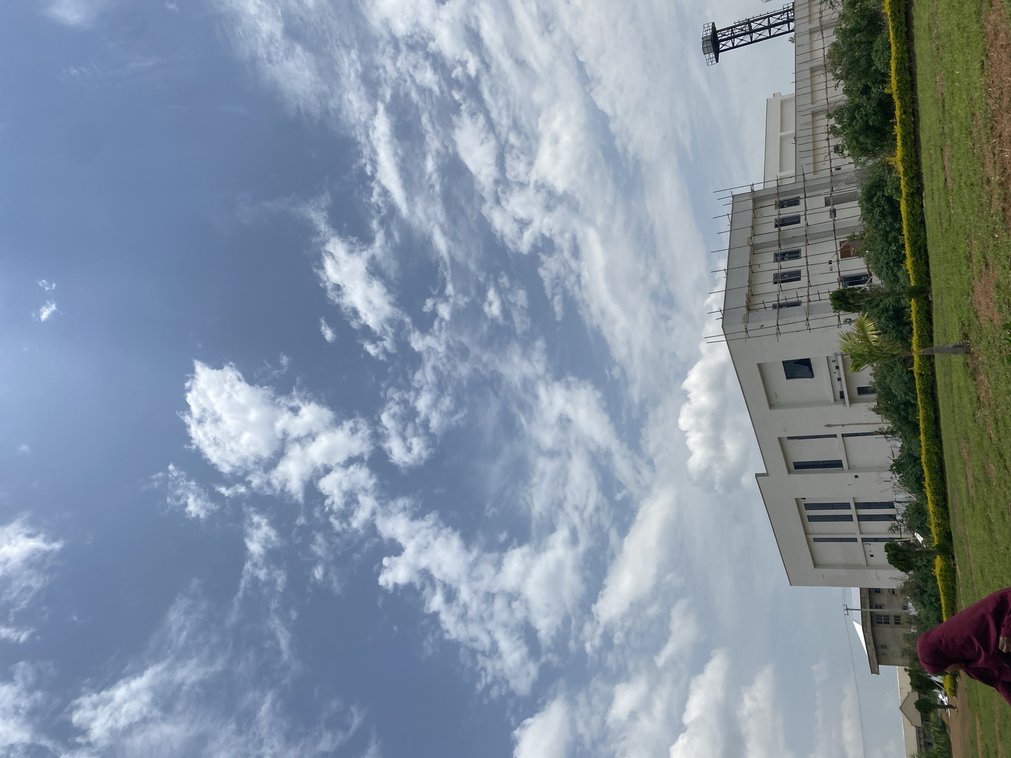 The image captures a building in Gwafan, Nigeria, under a sky with scattered clouds. The building is white, multi-storied, and appears to be undergoing some construction or maintenance as scaffolding is visible on one side. Greenery, likely a manicured hedge and some trees, lines the edge of the building, and a grassy area extends from it. A tall, thin tower is visible behind the building. The photo appears to have been taken from a low angle. In the lower right, a person can be seen, although obscured.