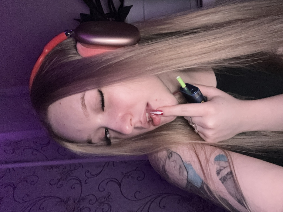 A female individual is depicted in a close-up, angled portrait, looking towards the camera with her left eye partially closed. She has long, straight blonde hair with darker roots, and is wearing rose gold over-ear headphones. Her right hand is raised, with the index finger, adorned with red and white nail polish, touching her slightly parted lower lip. Multiple rings are visible on her fingers. She holds a black device with a neon green tip, consistent with an e-cigarette or vape pen, in her right hand below her index finger. Visible on her left upper arm is a tattoo featuring a female face in blue ink. She is wearing a dark-colored top. The background is a dimly lit, dark purple wall with a subtle patterned design, and a dark green plant is partially visible behind her right shoulder. The specific location in Saratov, Russia, cannot be determined from the visual evidence provided in the image.