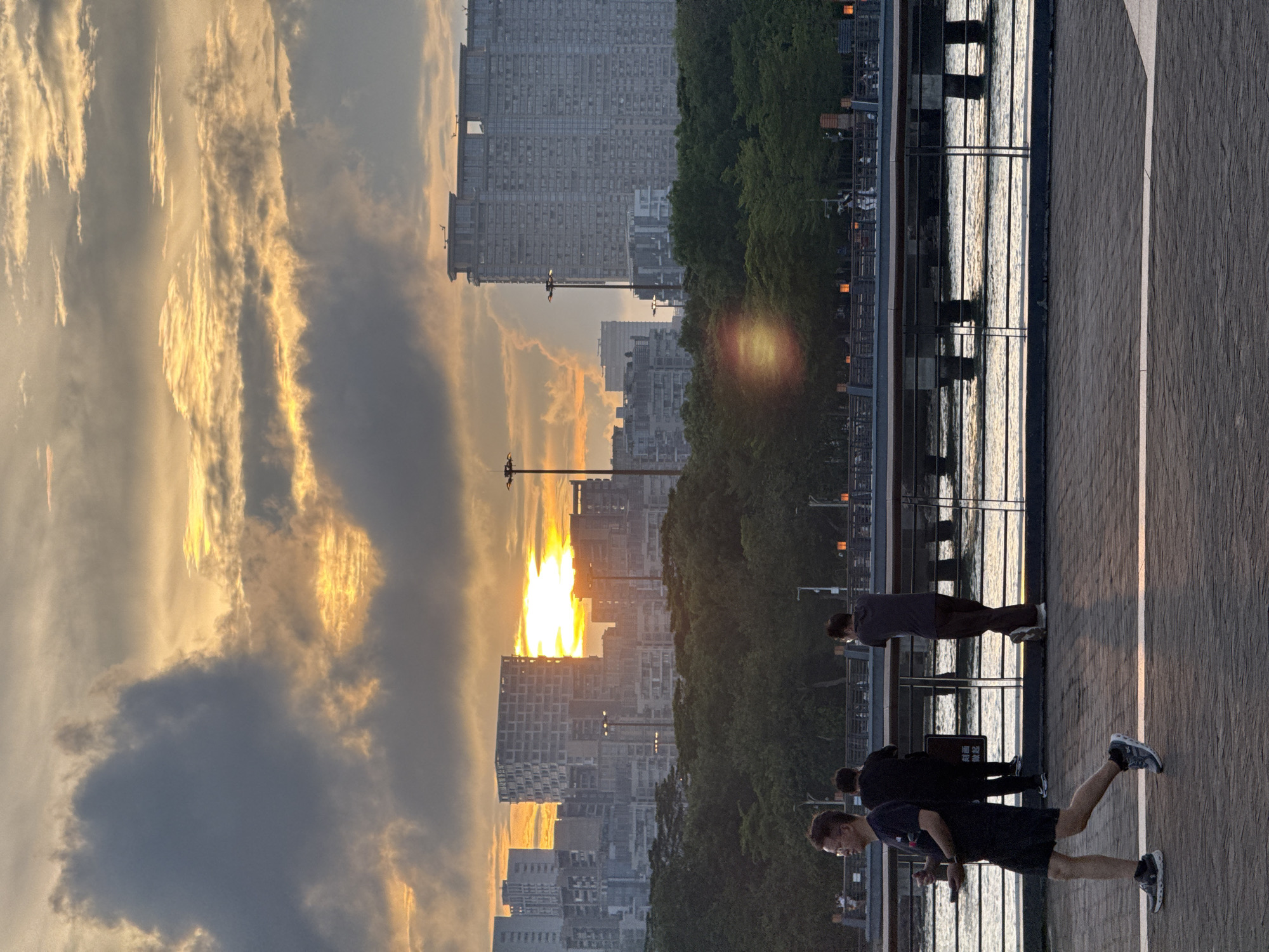 The image captures a sunset scene in the Nanshan District of Shenzhen, China. In the foreground, a pedestrian walkway runs along the waterfront, where a few people are present. One person is stretching or exercising, while others are walking or standing. A railing separates the walkway from the water. In the background, there is an urban landscape featuring high-rise buildings partially obscured by trees. The setting sun is visible behind the buildings, casting a golden glow on the clouds. The sky is filled with a mix of dark and illuminated clouds.