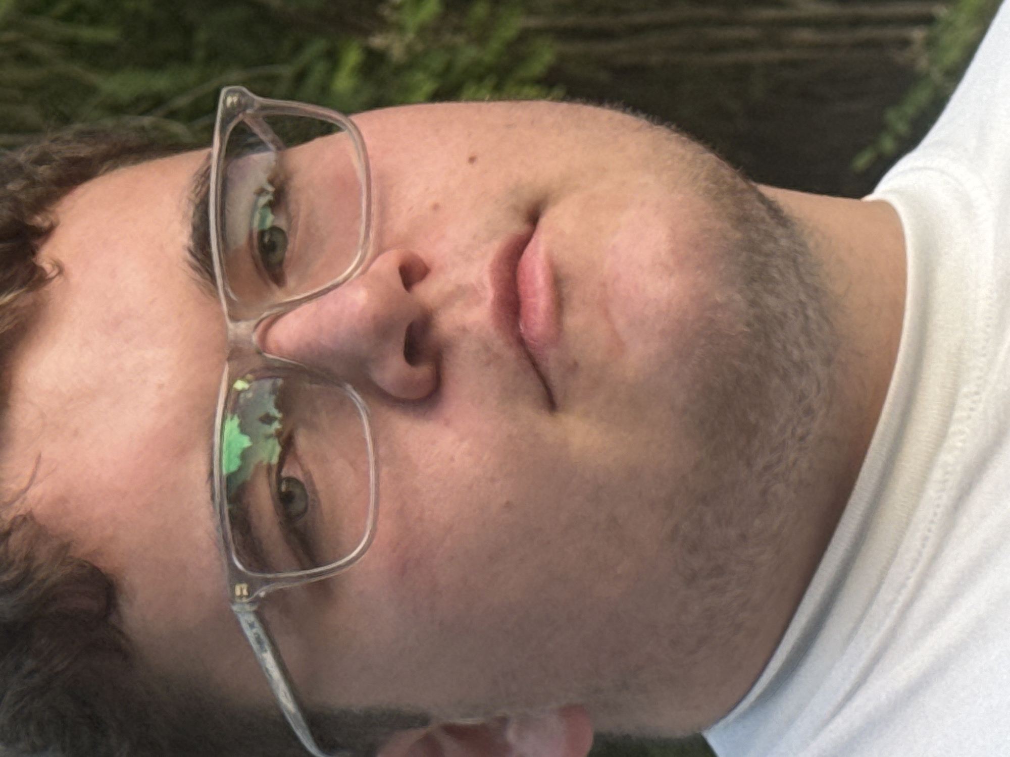 A close-up shot of a person wearing clear-framed glasses and a white T-shirt, positioned outdoors in front of a blurred green background. The person appears to be lying down, with their face angled towards the camera. The glasses reflect the surrounding environment, possibly foliage. There is no indication of the location being Pleven, Bulgaria.