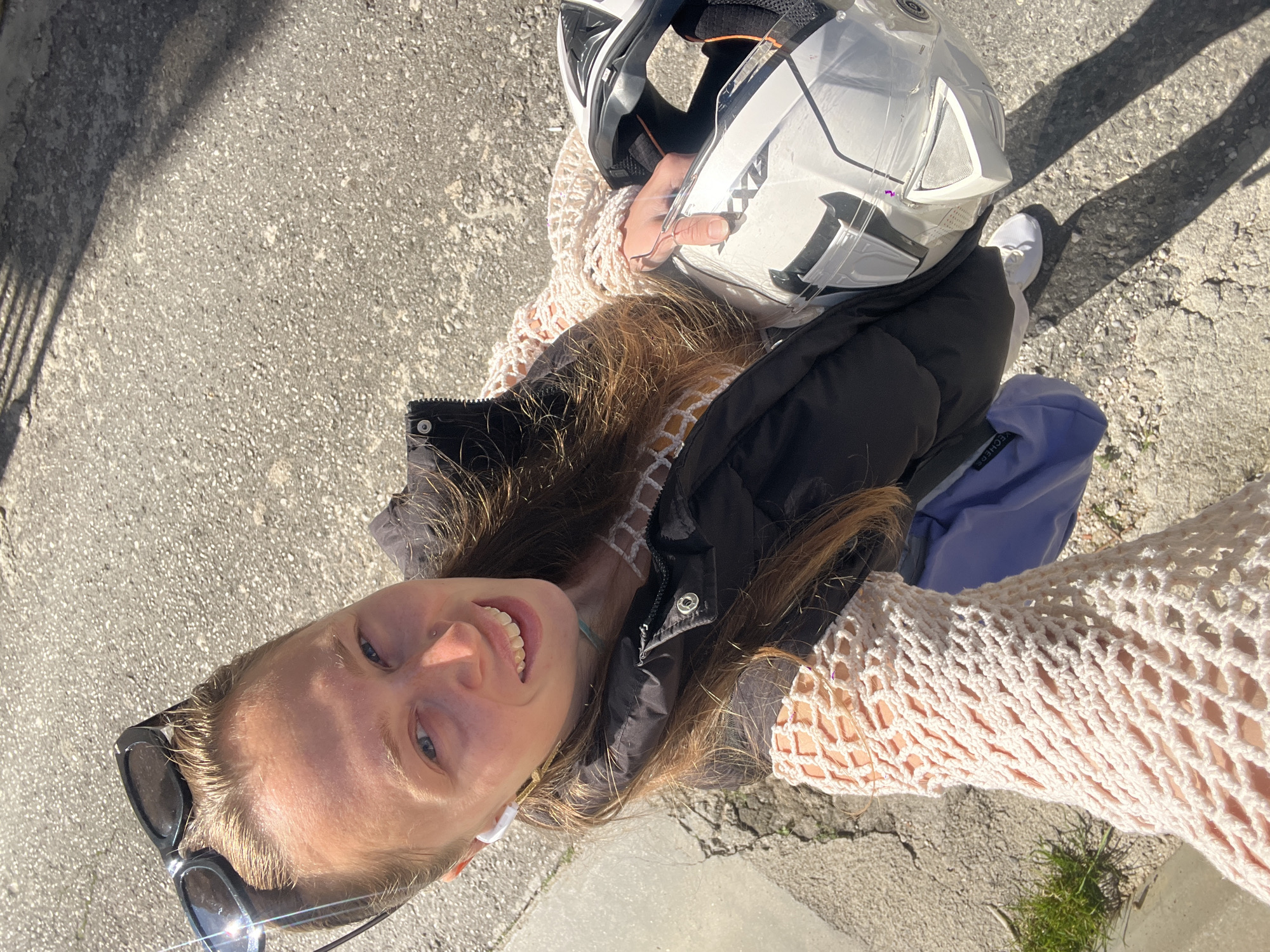 The image is an eye-level selfie taken outdoors on a sunny day. A young woman with light skin and long brown hair is in the foreground, smiling at the camera. She is wearing sunglasses pushed back on her head, a black vest, and a cream-colored crocheted shirt.
She is holding a white motorcycle helmet with black accents. Part of a white sneaker is also visible. A light blue bag is near her.
The background is a gray paved surface, indicating an urban or semi-urban area. Shadows are cast by the sun and objects in the scene. A patch of green vegetation is visible at the bottom edge of the frame. The location is likely Rafailovići, Montenegro, based on the nature of the scenery.