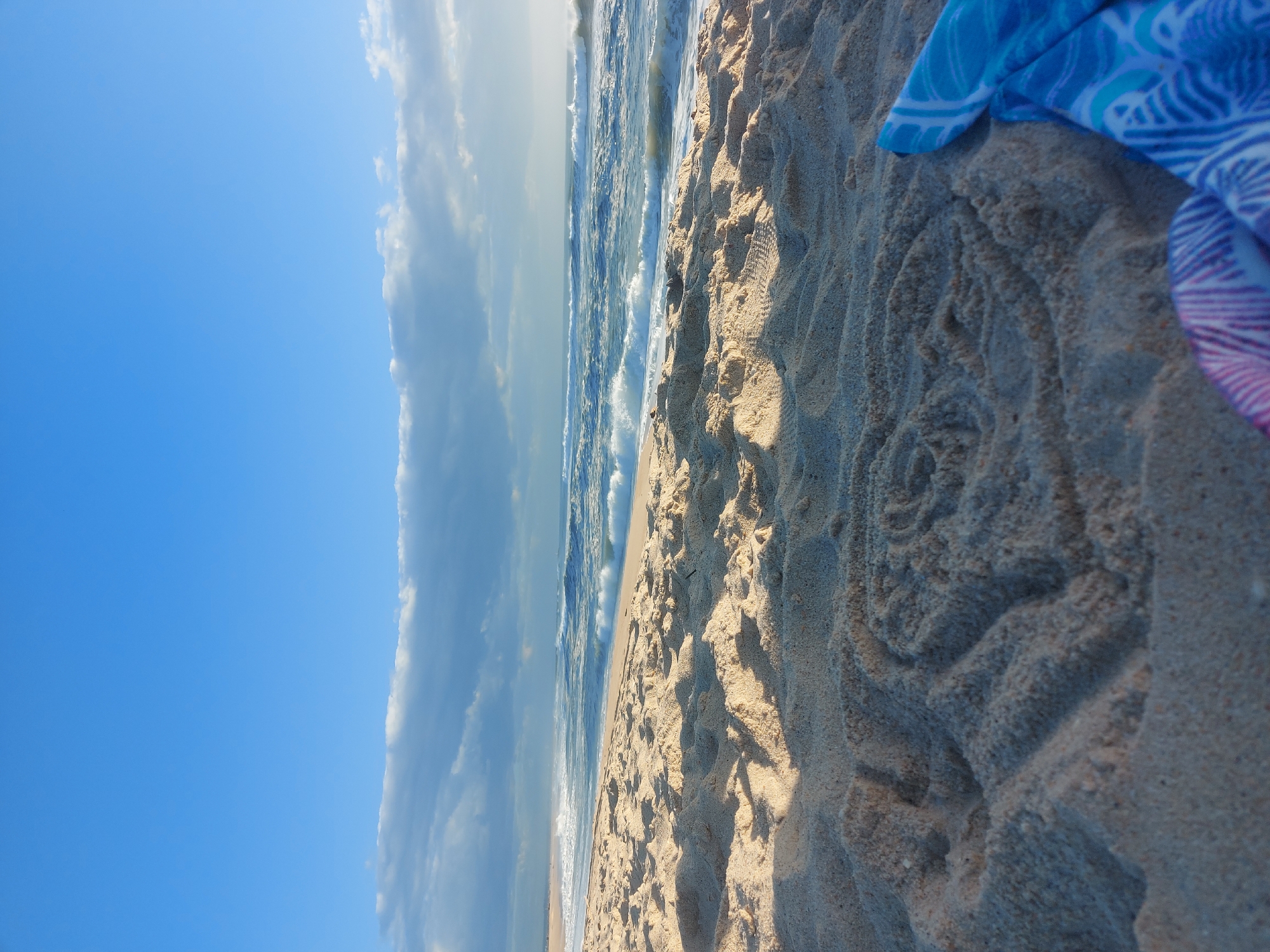The image depicts a beach scene in Figueira da Foz, Portugal. The perspective is ground-level, showing the sandy beach in the foreground. The sand exhibits patterns and footprints. To the left, the ocean meets the shoreline with visible waves. A blue-patterned towel is partially visible on the right side of the frame. The sky is clear blue with some cloud formations.