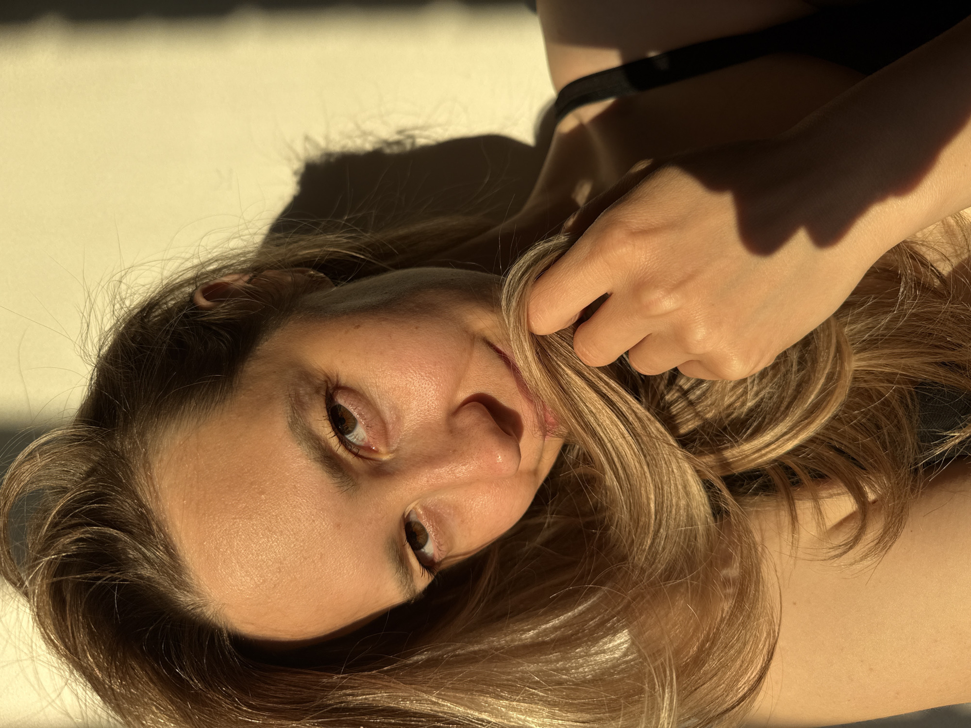 This close-up portrait features a young woman reclining, looking directly towards the viewer with a gentle, engaging expression. Her head is slightly tilted, and her long, wavy hair, a mix of light brown and blonde, cascades around her face and shoulders. Her right hand is gently placed in her hair near her cheek. She has light skin, expressive brown eyes, and a subtle smile. She appears to be wearing a dark top, with a strap visible on her right shoulder.
The scene is dominated by strong, warm, natural light, likely direct sunlight, which highlights her features and hair. This directional lighting casts prominent, elongated shadows across the light-colored, plain background, which could be a wall or floor. The quality of light and the long shadows suggest the photograph was taken during either early morning or late afternoon, often referred to as the "golden hour," indicating clear and sunny weather. The atmosphere conveyed is calm and intimate. There are no discernible objects beyond the subject, nor any visible text. While the location context is Chaikovsky, Russia, no specific geographical cues are visible within the image itself.