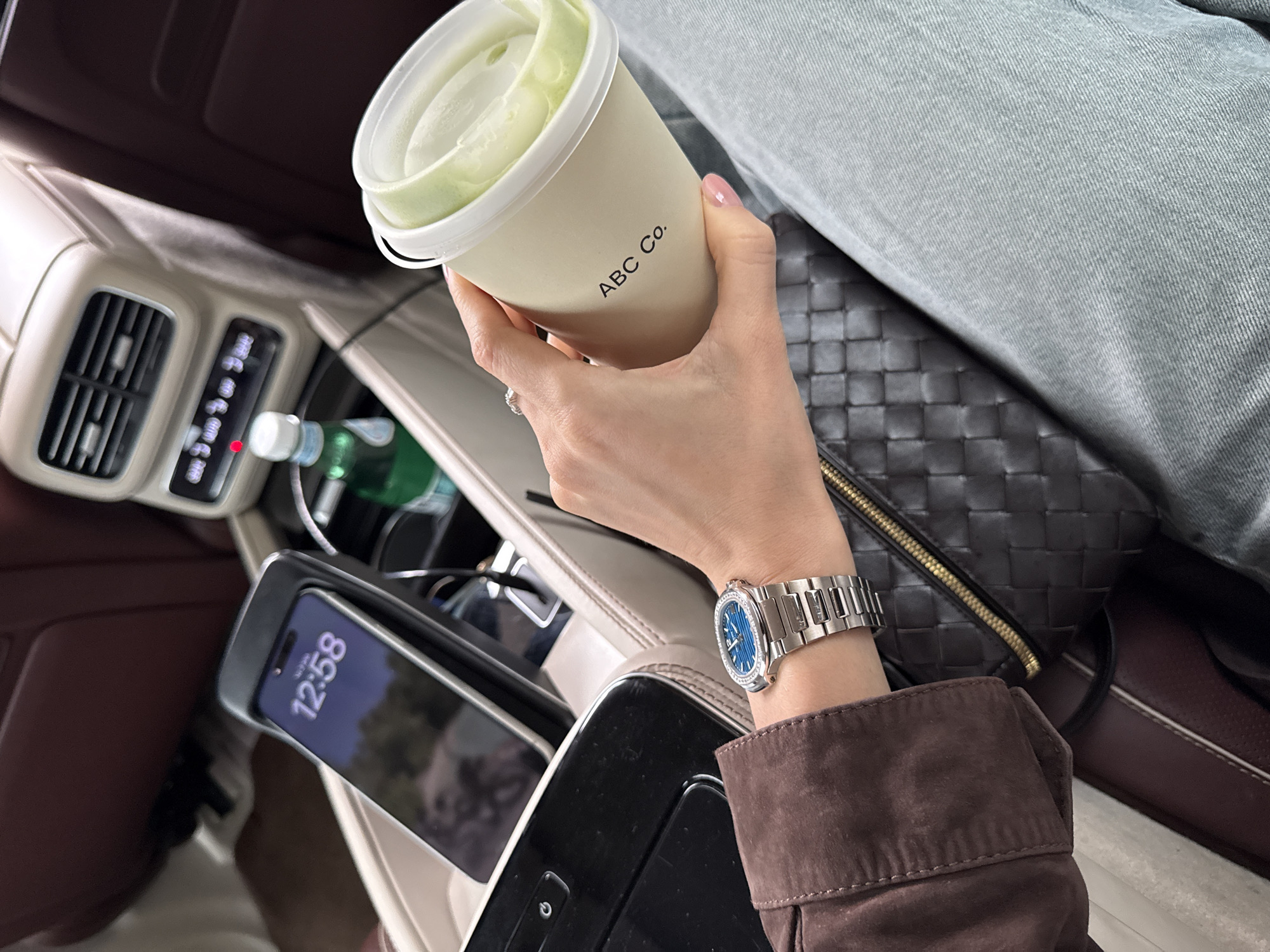 A high-angle, close-up shot inside a luxury car. A person is holding a cup with the text "ABC Co." printed on it, likely a beverage with a light green, foamy substance at the top. The person is wearing a silver wristwatch on their left wrist, and a brown jacket. Their other hand has a ring on its finger. In the car's console, a smartphone displays the time "12:58." A water bottle and other unidentified items are also visible. The background features parts of the car interior, including a vent and leather upholstery, and a woven black bag, with a part of a light gray pair of jeans as well. The setting lacks distinct landmarks or environmental cues to definitively determine it is in Moscow, Russia.