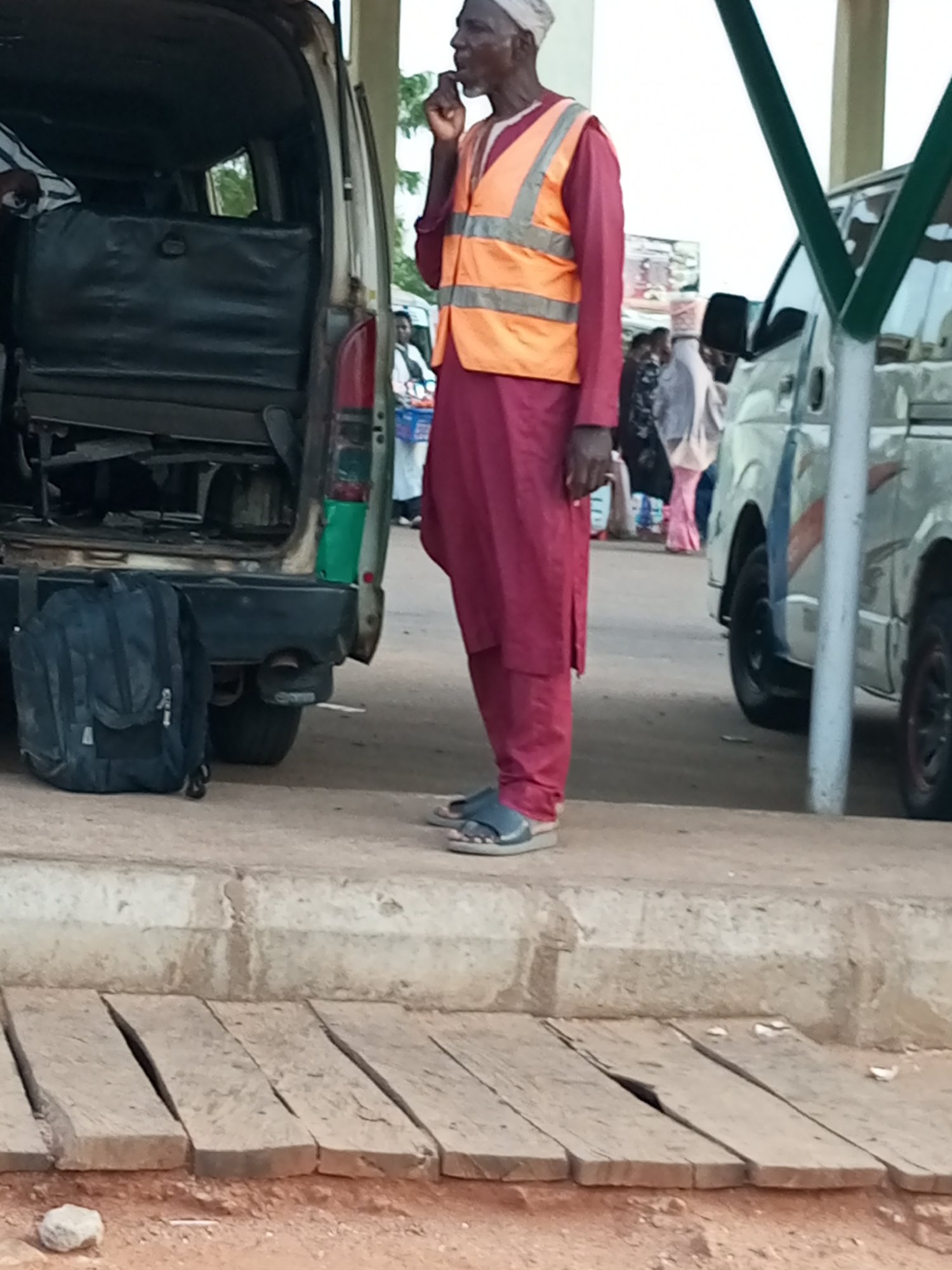 The image depicts a street scene in Gombe, Nigeria, with a man in a maroon traditional outfit and an orange safety vest standing next to a white van. He is wearing a white cap and sandals. A black backpack is visible on the ground near the rear of the van. In the background, other people and vehicles are present, suggesting a busy public area. The environment appears to be an outdoor setting with a paved ground and a curb. The lighting suggests it is daytime.
