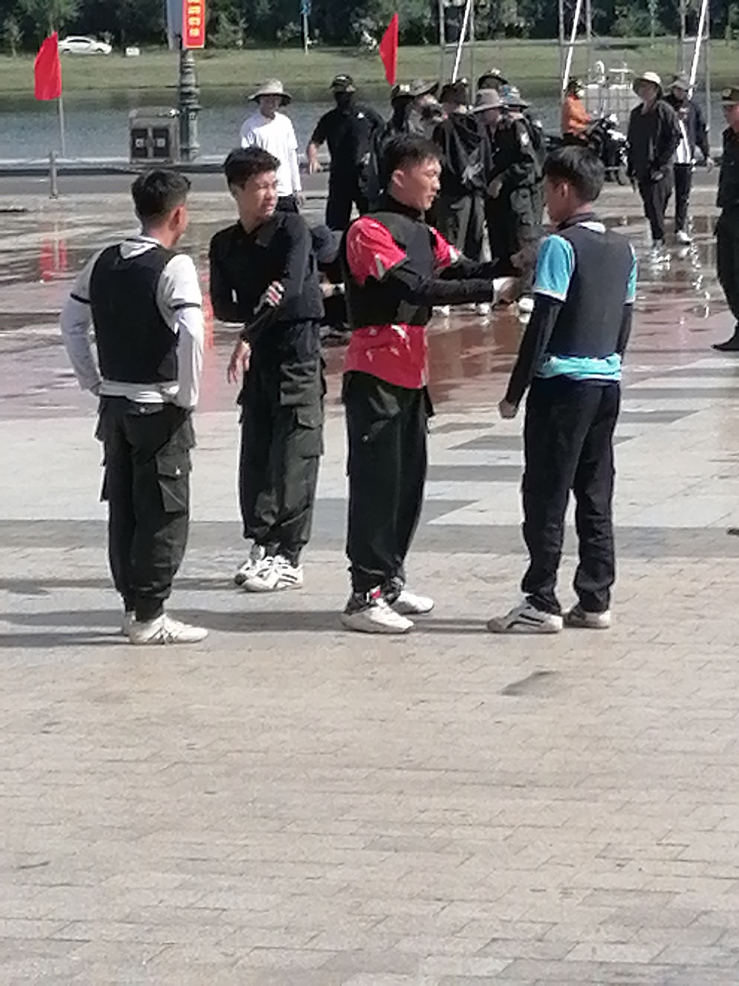 The image depicts a group of young men in athletic wear gathered on a paved, light-colored, patterned ground that appears damp, suggesting recent rain or a water feature. The background shows a body of water, a low barrier fence, trees, and a building. There are also several other people in the background, some wearing hats.
In the foreground, four young men are the primary subjects. From left to right:
1. **Man 1:** Wearing a white long-sleeved shirt, a black vest, and black cargo pants. He has white athletic shoes. He is standing with his hands behind his back, looking towards the man to his right.
2. **Man 2:** Wearing a black long-sleeved shirt and black cargo pants. He is looking towards Man 1.
3. **Man 3:** Wearing a red and black jacket with black sleeves, and black pants. He is gesturing with his right hand towards Man 4, who is to his right.
4. **Man 4:** Wearing a blue and black t-shirt, a black vest, and black pants. He has a light blue sash or band around his waist. He is wearing white and grey athletic shoes and is looking towards Man 3.
The setting appears to be an outdoor public space, possibly a plaza or park, with elements of a waterfront. The presence of athletic attire and the setting might suggest a sports-related event or training. The location in Da Lat, Vietnam, is not visually evident from the provided image.