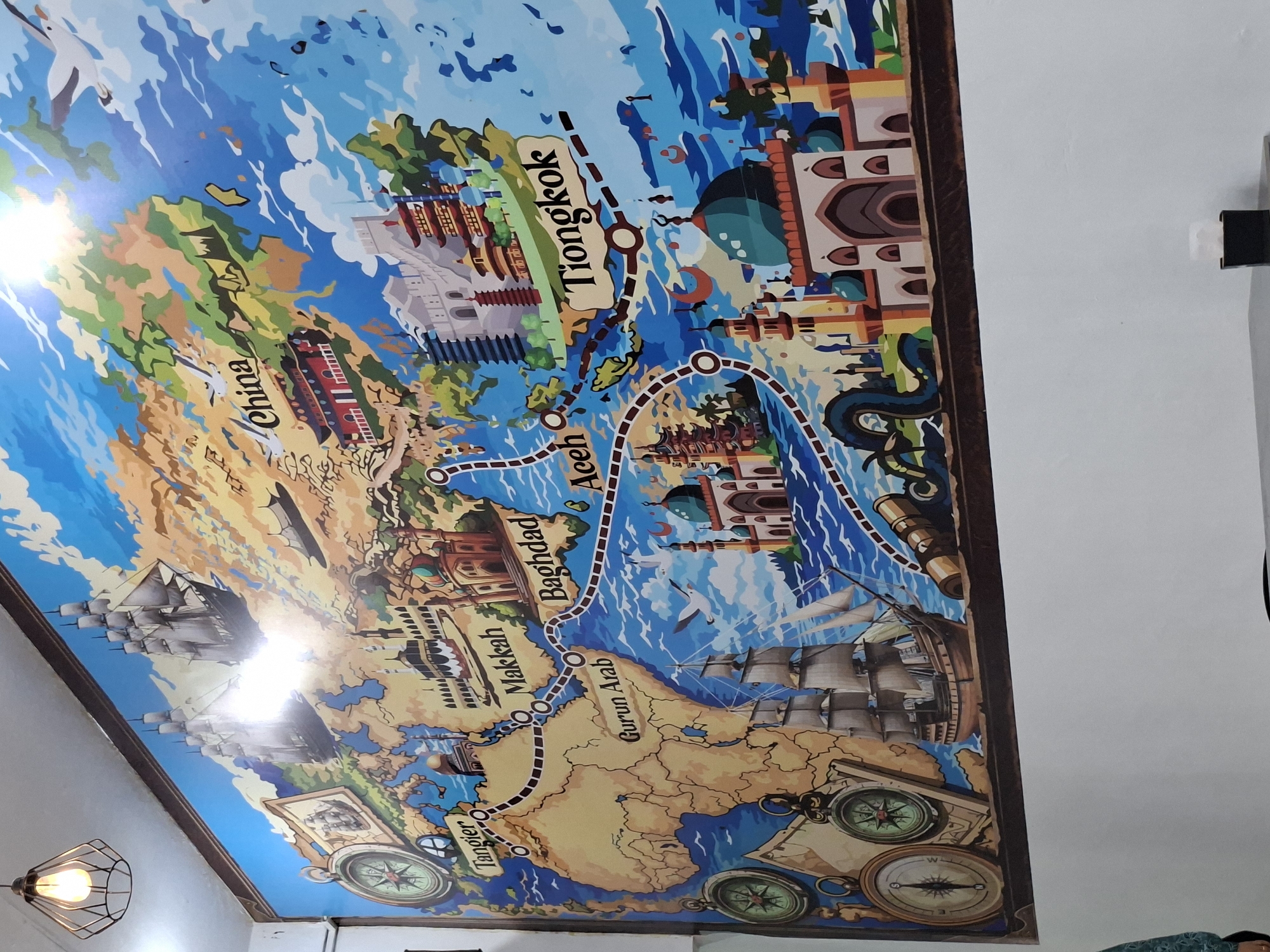 The image shows a decorated ceiling or wall panel featuring a stylized historical world map. The map depicts various landmarks and cities connected by a dotted route, suggesting a journey. Cities like "Tiongkok," "Aceh," "Baghdad," "Makkah," and "Tangier" are labeled, indicating locations across Asia, the Middle East, and North Africa. The artistic style includes illustrations of ships, compasses, and historical buildings associated with these regions. The environment is an interior space, as indicated by the white wall and a light fixture visible on the lower left side of the picture. The location in the city of Lhokseumawe, Indonesia, is not visually verifiable from the image alone; it might be the location of where the photograph was taken. There are no people visible in the image.