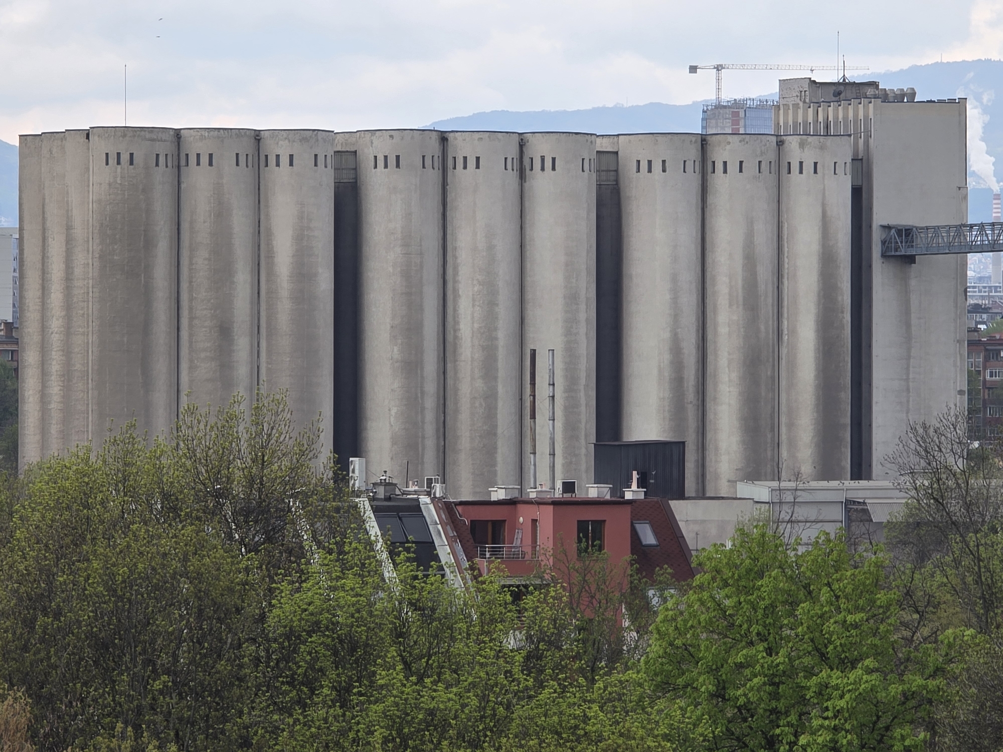 The image displays a panoramic view of a large industrial complex dominated by a row of massive, cylindrical concrete silos, likely grain elevators, situated in Sofia, Bulgaria. These tall, uniformly gray structures feature numerous small, rectangular openings near their tops and distinct vertical seams. On the far right, the silos merge with a taller, rectangular concrete building, which is connected by a metal truss bridge, suggesting a conveyor system.
In the mid-ground and foreground, a dense canopy of lush green trees, indicative of spring or early summer foliage, partially obscures the lower parts of the industrial site and an adjacent residential area. Nestled among these trees is a red-roofed building with several windows and a small balcony, showing signs of residential or commercial use, including visible pipes, vents, and air conditioning units on its roof. Other less distinct buildings are visible peeking through the trees.
The background reveals a range of hazy mountains under an overcast, cloudy sky. A large construction crane is visible in the distance, hinting at ongoing development, and faint industrial smoke plumes can be seen further right. A few tiny bird specks are scattered across the sky. The lighting is diffused and soft, characteristic of an overcast day. No discernible text is visible on any of the structures. The scene presents a juxtaposition of heavy industry with urban and natural elements.