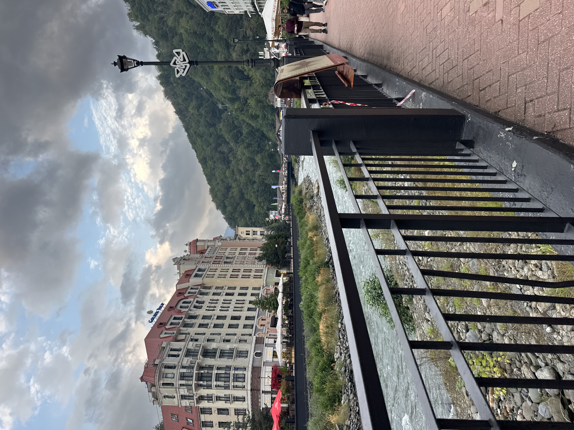 The image shows a paved walkway alongside a river in Estosadok, Russia. A black metal railing with vertical bars runs along the edge of the walkway, separating it from the river. The river is flowing and appears to have rocky banks. Across the river, a large, multi-story building with a red-tiled roof and white facades stands prominently. This building features numerous windows and balconies. Beyond this building, a lush green mountain slope rises, covered in trees. The sky above is filled with dramatic, overcast clouds, with patches of lighter sky visible. The walkway is made of red brick pavers laid in a herringbone pattern. A black lamppost with an ornate decorative element is visible on the right side of the walkway. Two individuals are walking away from the camera on the walkway. One is wearing a burgundy sweater and khaki pants, and the other is wearing dark clothing. A brown framed object, possibly a menu or a notice board, is leaning against the railing. The overall scene depicts a scenic, possibly resort, area with a blend of urban architecture and natural mountainous landscape.