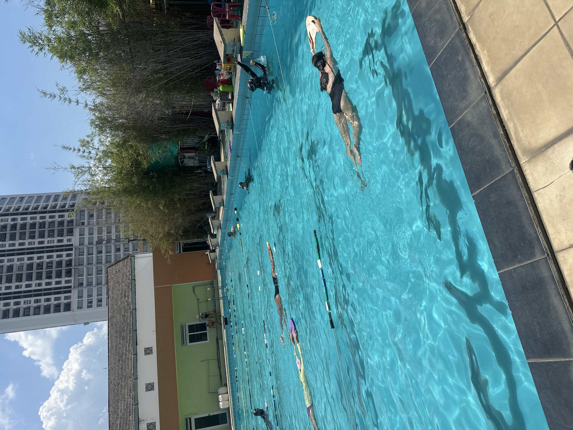 The image shows an outdoor swimming pool, likely a public or club facility, located in Nha Trang Ward, Vietnam, as evidenced by the local architecture and foliage. The pool is a rectangular shape with multiple lanes, indicated by lane markers. Several people are in the pool, swimming in various lanes. One individual is using a kickboard, while another appears to be diving or starting from the edge. The pool is surrounded by tiled decking and a building with green, orange, and white sections. Bamboo trees line one side of the pool. In the background, a tall apartment building is visible against a blue sky with white clouds. The lighting suggests it is a sunny day.