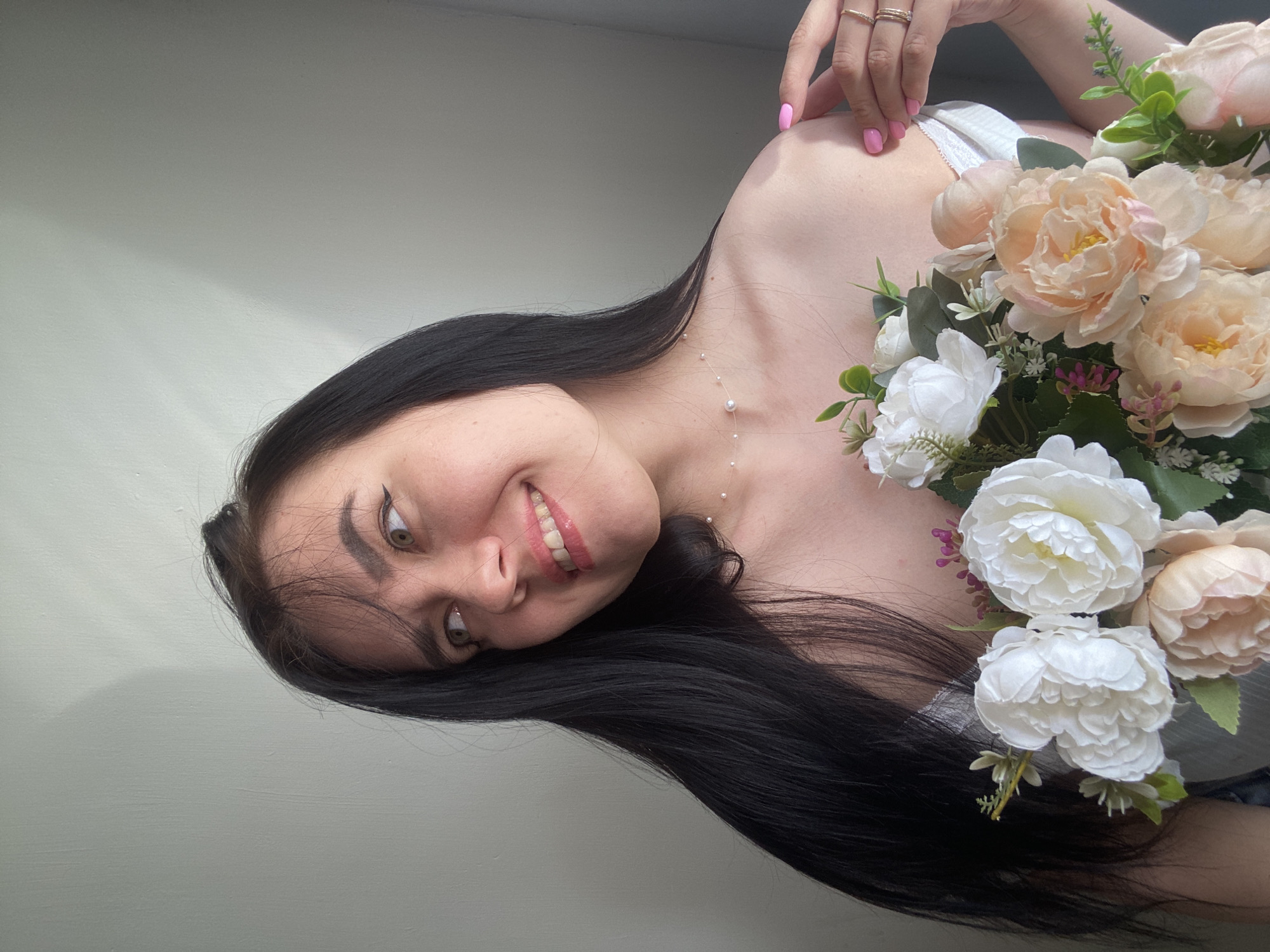 The image shows a young woman with long, dark hair and light eyes, holding a bouquet of artificial flowers. She is wearing a white off-the-shoulder top and a delicate pearl necklace. Her right hand, with bright pink fingernails and rings, is resting on her left shoulder. The background is a plain, light-colored wall with a shadow on the left side. The woman is smiling and looking to her left. The scene appears to be indoors, possibly in a residential setting in the city of Chaikovsky, Russia.