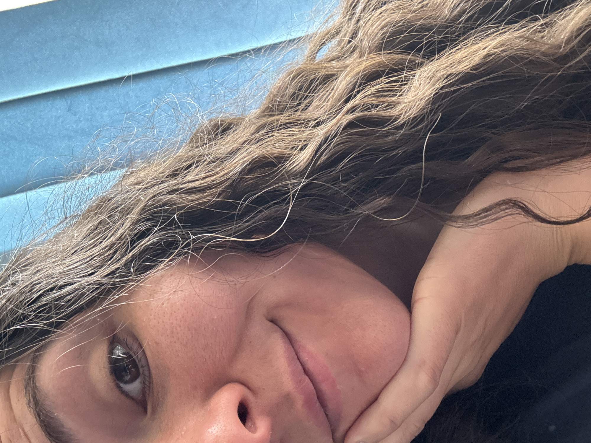 A close-up, slightly angled image shows a person with light brown, wavy hair and fair skin. The person's right eye is visible, along with their nose and mouth, which is slightly puckered. Their right hand is positioned against their cheek, suggesting they are resting their face in their hand. The background includes a light blue, possibly textured surface. The specific location in the city of Unknown, Unknown is not discernible from the image.