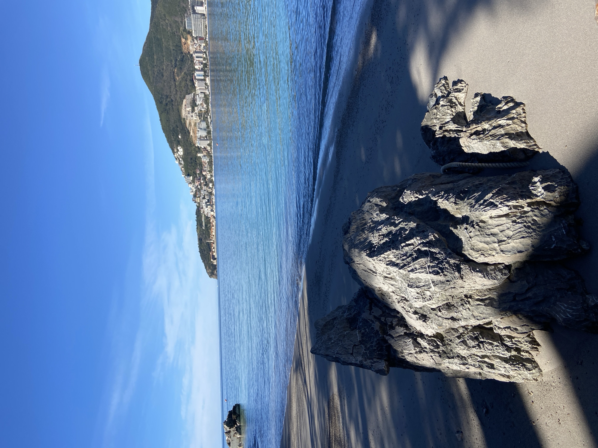 The image shows a beach in Budva, Montenegro on a sunny day. The beach is sandy with a large rock formation in the foreground. The sea is calm and blue, with a gentle wave lapping on the shore. In the background is a mountain with buildings scattered along its slope, indicating a built-up area. The sky is clear blue with faint wisps of clouds. The image is rotated, with the sky on the left side.
