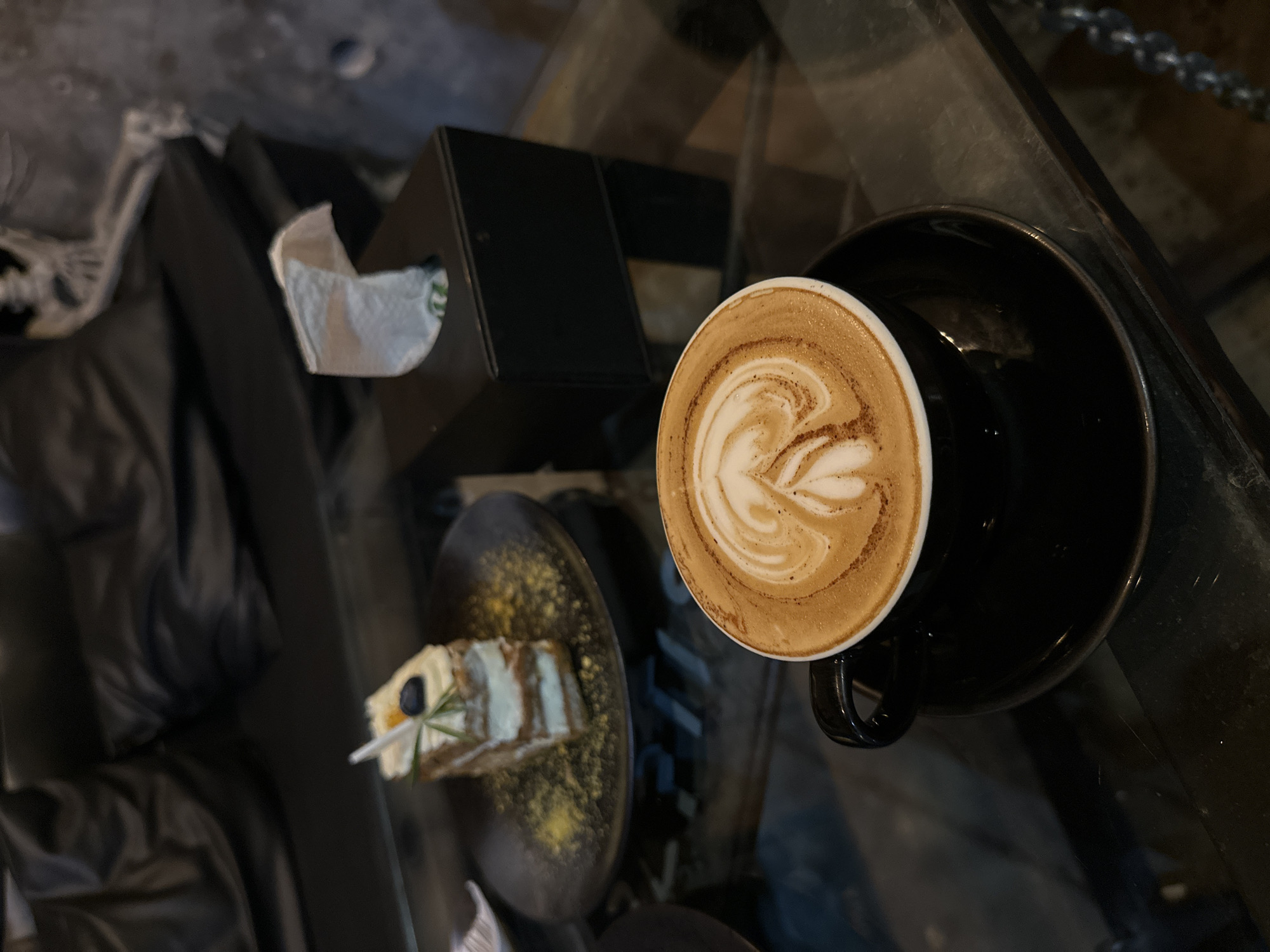 In a dimly lit, modern cafe, likely in Phuket City Municipality, Thailand, a slice of layered cake with white frosting, a blueberry, and a sprig of rosemary sits on a black plate dusted with yellow powder. Nearby, a black tissue box dispenses a crumpled white tissue. The focal point is a black mug filled with frothy latte art, placed on a matching saucer. The table is made of reflective, dark glass, with the blurred background revealing black leather seating.