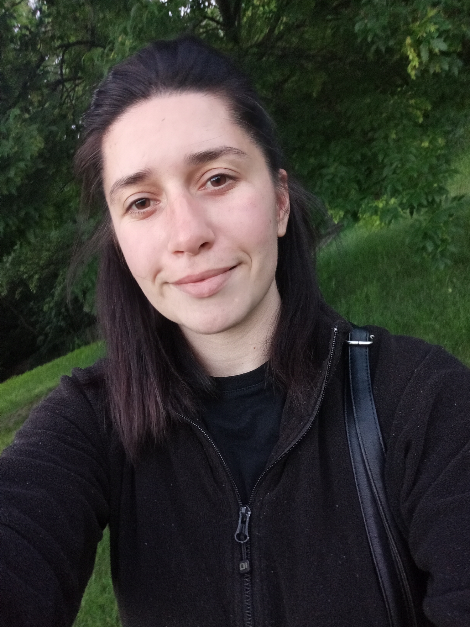 A close-up, outdoor selfie captures a young woman from the chest up. She appears to be in her 20s or 30s, with dark, shoulder-length hair pulled back from her face, revealing a clear forehead. Her dark brown eyes gaze directly at the camera, and she wears a subtle, gentle smile with closed lips. Her complexion is fair.
She is dressed in a dark, likely black, zip-up fleece jacket, worn over a dark t-shirt. The texture of the fleece is visible, appearing soft and slightly fuzzy. A black shoulder strap from a bag runs over her right shoulder. The metallic zipper pull on her jacket features a small, rectangular emblem with what appears to be a faint "DI" or similar geometric symbol engraved on it.
The background is softly blurred, consisting of lush green foliage from trees and a grassy slope, indicating a natural setting such as a park or forest edge. The lighting is soft and natural, suggesting it might be late afternoon or early evening. The overall atmosphere conveyed is calm and casual. The provided context indicates the location is Sofia, Bulgaria.