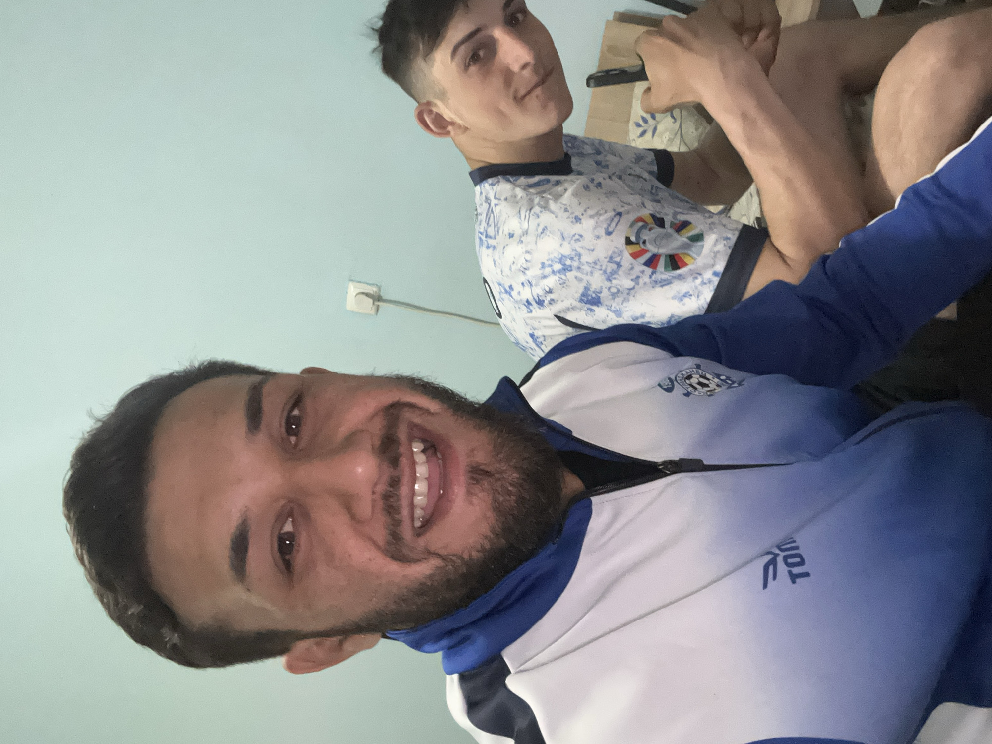 Two men are posing for a photo. The man in the foreground is smiling broadly, wearing a white and blue jacket. The man in the background is looking at the camera with a slight smirk, wearing a white and blue shirt. A Walmart bag is visible behind him. The background is a light blue wall with an electrical outlet. There is no information about the location in Tsarevo, Bulgaria.