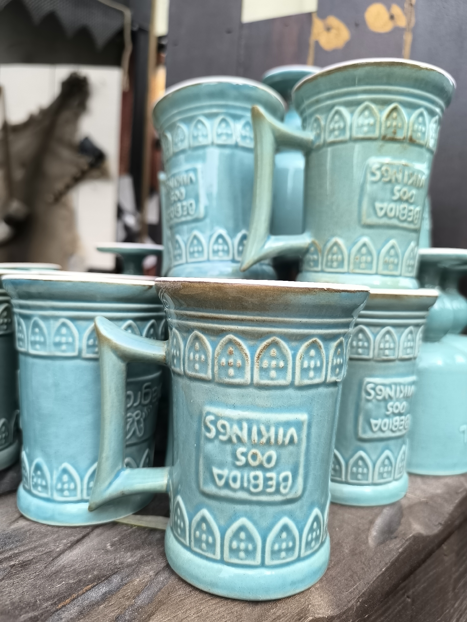 A close-up shot showcases a collection of turquoise mugs stacked on a wooden surface. The mugs are ornately decorated with embossed patterns, featuring a band of arched motifs near the rim and a rectangular panel with raised lettering. The text on the visible mugs reads "BEBIDA DOS VIKINGS" and "500" in reverse. One mug, positioned prominently in the foreground, is angled to reveal its handle and a partial inscription. The background is blurred, but hints of a shadowy figure and what appears to be a display rack suggest an outdoor market setting. The mugs are likely ceramic and exhibit a slightly aged appearance with subtle variations in color and texture. The scene is likely located in Óbidos, Portugal, a city known for its historic charm and artisanal crafts, and it is probable that these mugs are a souvenir item sold within the city.