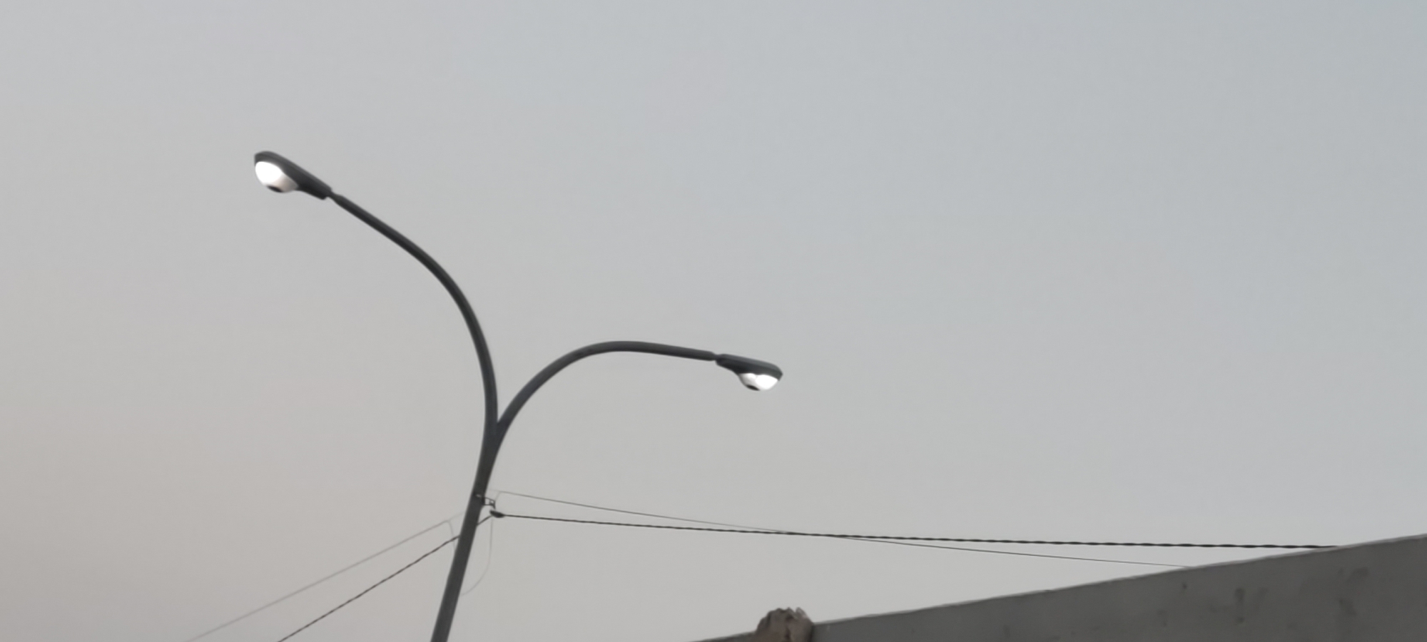 The image shows two streetlights against a pale gray sky, suggesting an overcast day or dusk. The streetlights are modern in design, with dark, curved poles. The one on the left is taller and arcs over to the right, with its light emitting a bright white glow. The one on the right is shorter and arcs to the right as well, also emitting a bright white light. Below the streetlights, a dark, thin wire is visible, strung between two unseen points. Further down, a light-colored, horizontal structure, possibly a wall or rooftop, occupies the bottom portion of the image. The overall scene is devoid of any people or specific actions, presenting a still, functional urban landscape. This scene is located in Pakusamben, Indonesia.
