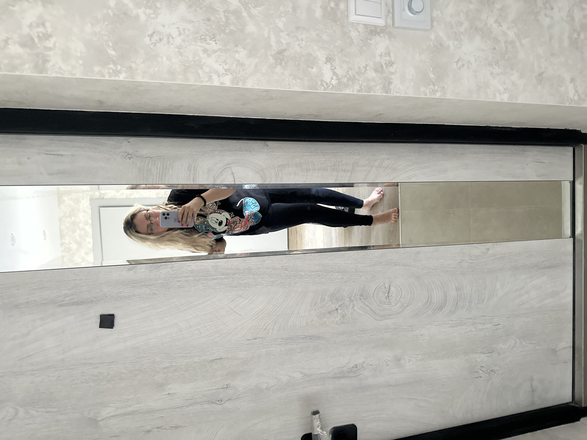 A woman is reflected in a full-length mirror, taking a selfie with her phone. She is wearing a black t-shirt with a colorful Mickey Mouse graphic and black leggings. She is barefoot and has blonde hair and glasses. The mirror is set against a wall with a light wood-grain finish and a dark vertical frame. To the left of the mirror is a light-colored door with a black handle. The floor is tiled with beige squares. The background of the reflection shows a room with light-colored walls and ceiling. The scene is indoors, likely in a residential setting, and is located in Novoaltaysk, Russia.