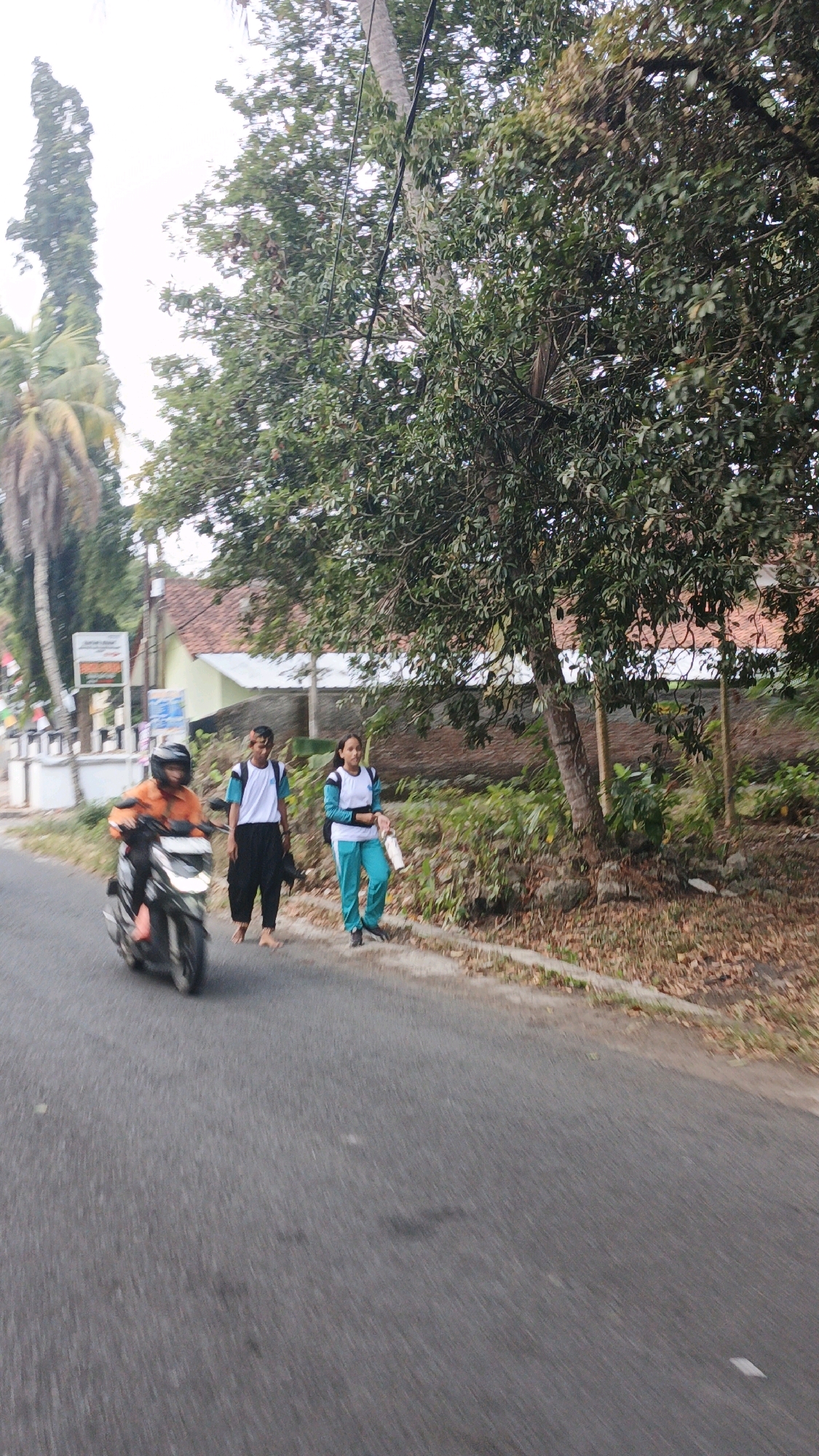 In Desa Karangjaladri, Indonesia, a scooter rider wearing an orange jacket and black helmet is on the left side of a paved road. Two pedestrians, a male in black pants and a white shirt with blue trim, and a female in teal pants and a white shirt with blue trim, are walking on the right side of the road. They appear to be students, possibly carrying bags. The environment features lush green trees, dry leaves on the ground, and a house with a red tile roof in the background. Power lines are visible overhead. The road is a typical rural Indonesian street.