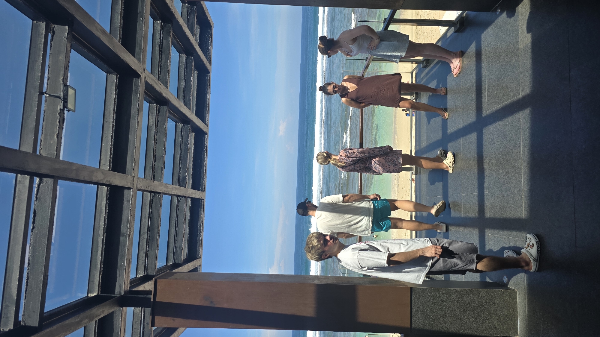 The image shows a group of five people standing on a balcony overlooking a beach. The location appears to be in Kuta Selatan, Indonesia, indicated by the beach, the ocean, and the architectural style. A structural grid partially frames the left side of the view. The people are casually dressed, in summer attire, some wearing dresses and others in shorts and t-shirts. The weather is sunny, with a blue sky and the ocean stretching to the horizon. White waves are visible along the shoreline. The balcony surface is dark.