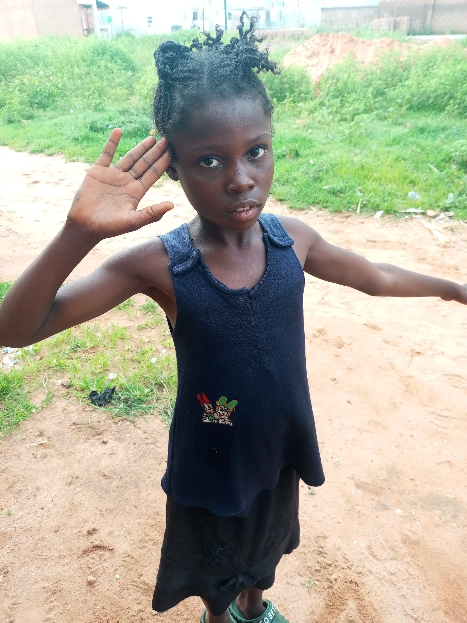 A young girl with dark skin and braided hair stands on a dirt path in Gombe, Nigeria. She is wearing a dark blue sleeveless top over a black skirt. Her right hand is raised to her forehead as if in a salute, and her gaze is directed forward. The background consists of tall green grass and some buildings. The ground is sandy and uneven, with some scattered debris.
