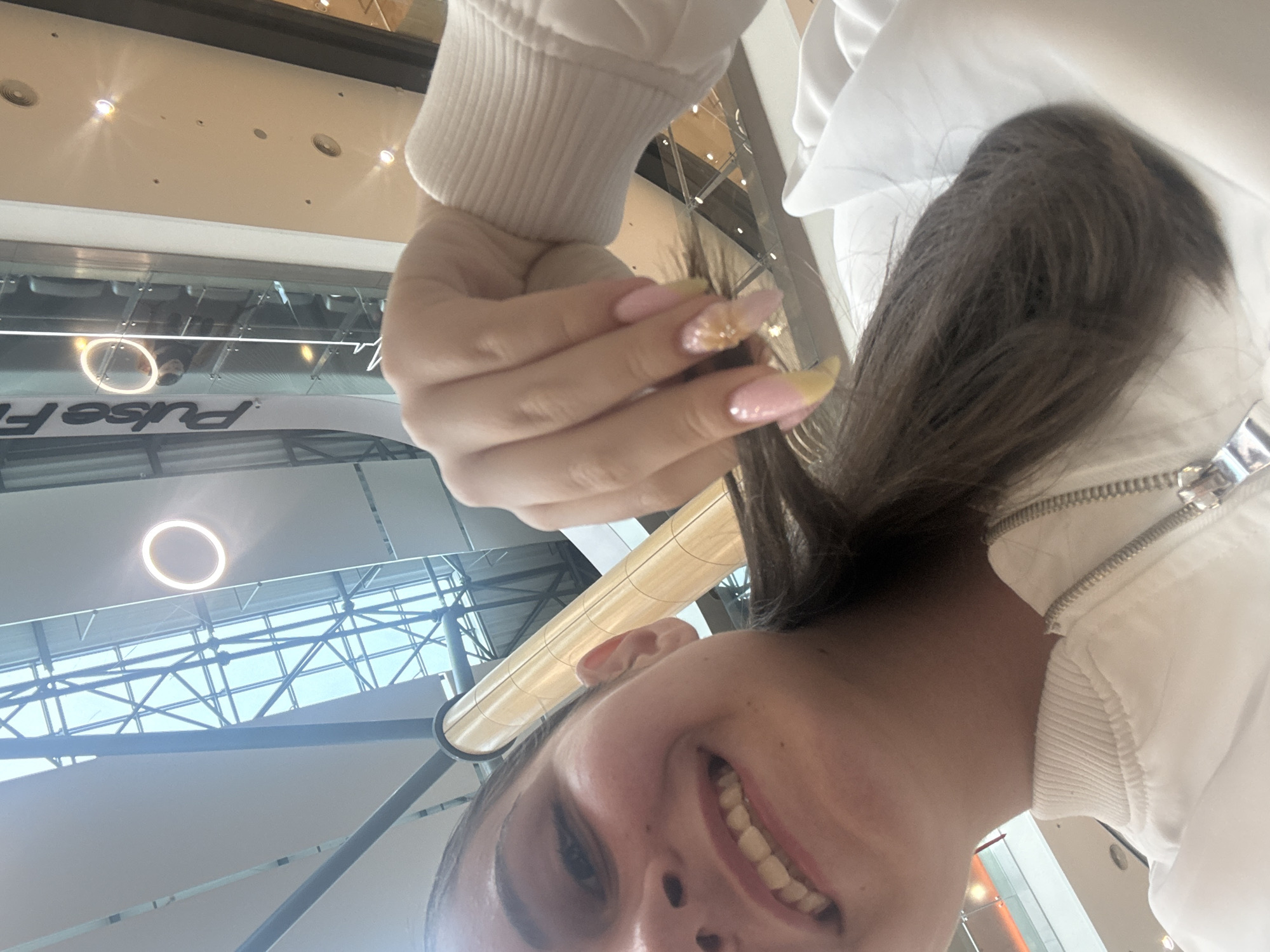 A selfie shot from a low angle depicts a young woman with brown hair pulled back, a white jacket, and manicured nails. She is smiling, and the camera is close to her face, capturing her features in detail. The background reveals an indoor space with a high ceiling, possibly a shopping mall or airport. A metal structure with crisscrossing beams is visible, along with circular light fixtures and signage that reads "Pulse F". The location is likely within the city of Burgas, Bulgaria, though precise confirmation would require more specific details from the signage.