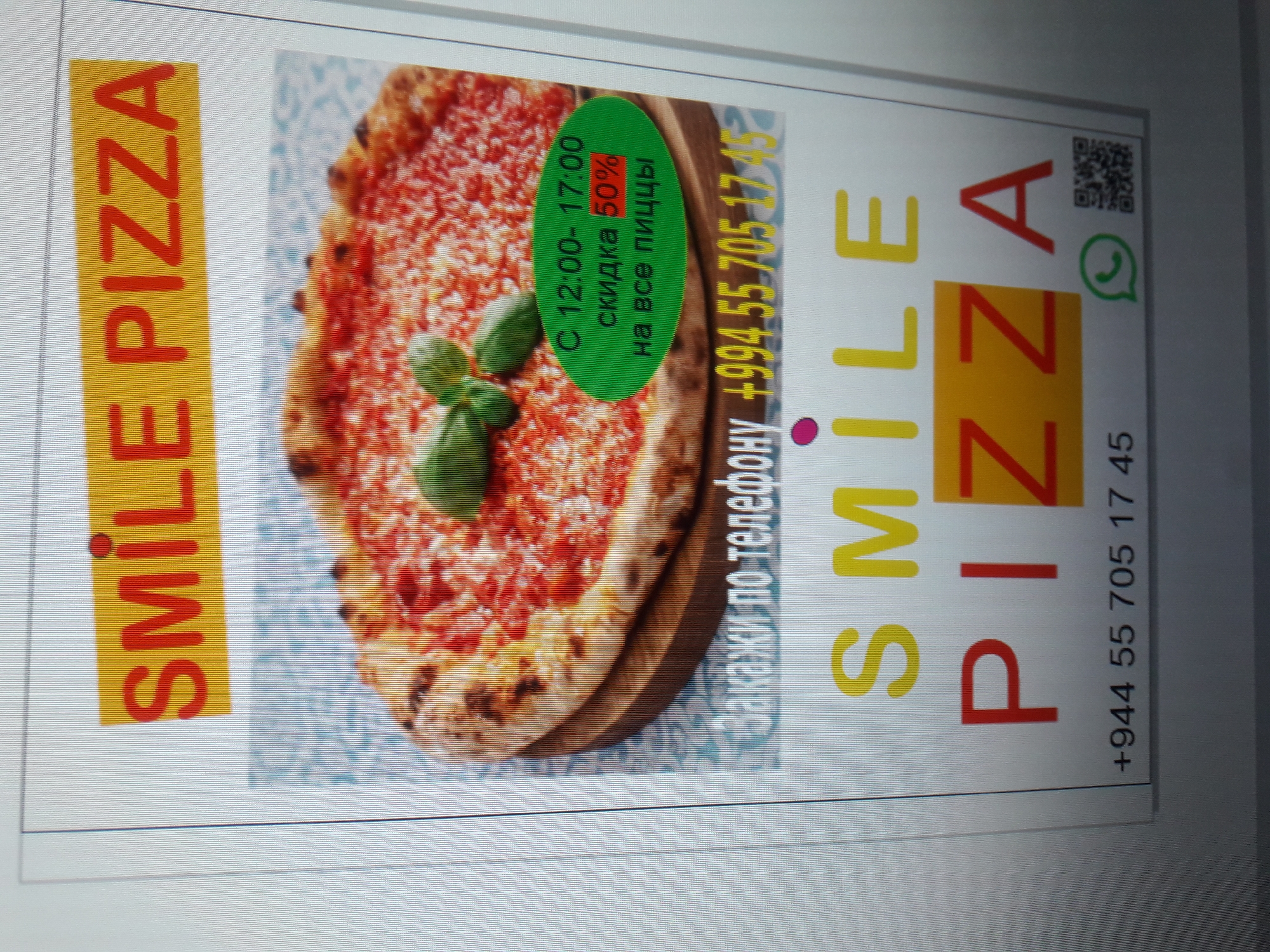 This is a digital image of a flyer advertising "SMILE PIZZA". The flyer features a large, appealing photograph of a pizza with fresh basil. Above the pizza, the text "SMILE PIZZA" is prominently displayed in red lettering against a yellow background. Below the pizza, there is a green oval shape containing text that reads "С 12:00- 17:00 скидка 50% на все пиццы", indicating a discount from 12:00 to 17:00 on all pizzas. Further down, the text "Закажи по телефону" is followed by a phone number "+994 55 705 17 45". The bottom of the flyer reiterates the brand name "SMILE PIZZA" in large, colorful letters. To the right of the phone number, there are icons for WhatsApp and a QR code. The overall presentation suggests a promotional advertisement for a pizza business. The image is captured from a computer screen, showing digital lines and pixels.