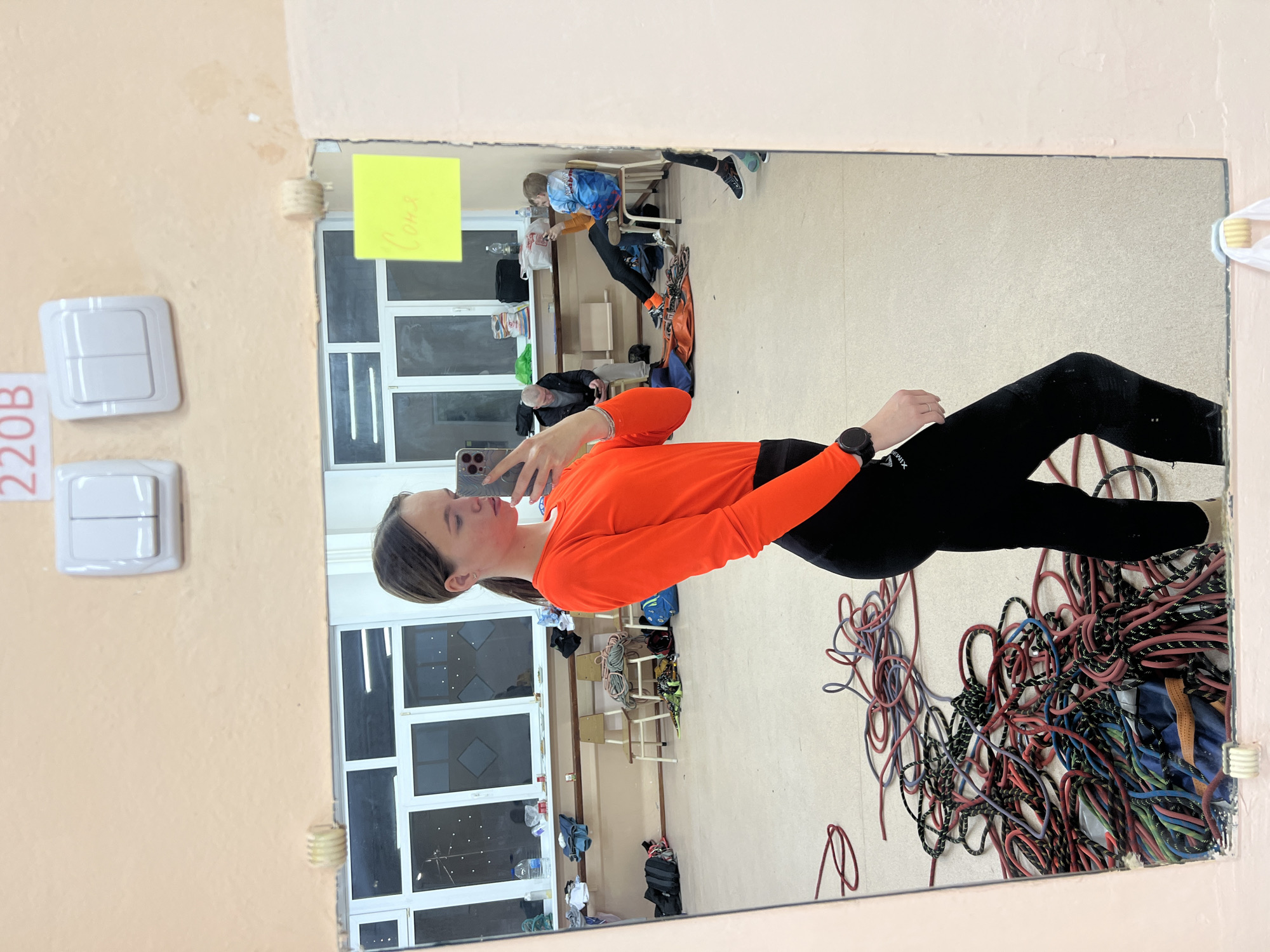 A young woman stands in front of a mirror, taking a selfie. She is wearing a long-sleeved orange shirt and black leggings. The reflection in the mirror shows a room with chairs, ropes, and a window. A sign with "Соня" written on it is taped to the mirror. The room appears to be in a school or training facility, given the presence of training ropes. Based on the information available, this image likely originates from Berezniki, Russia.