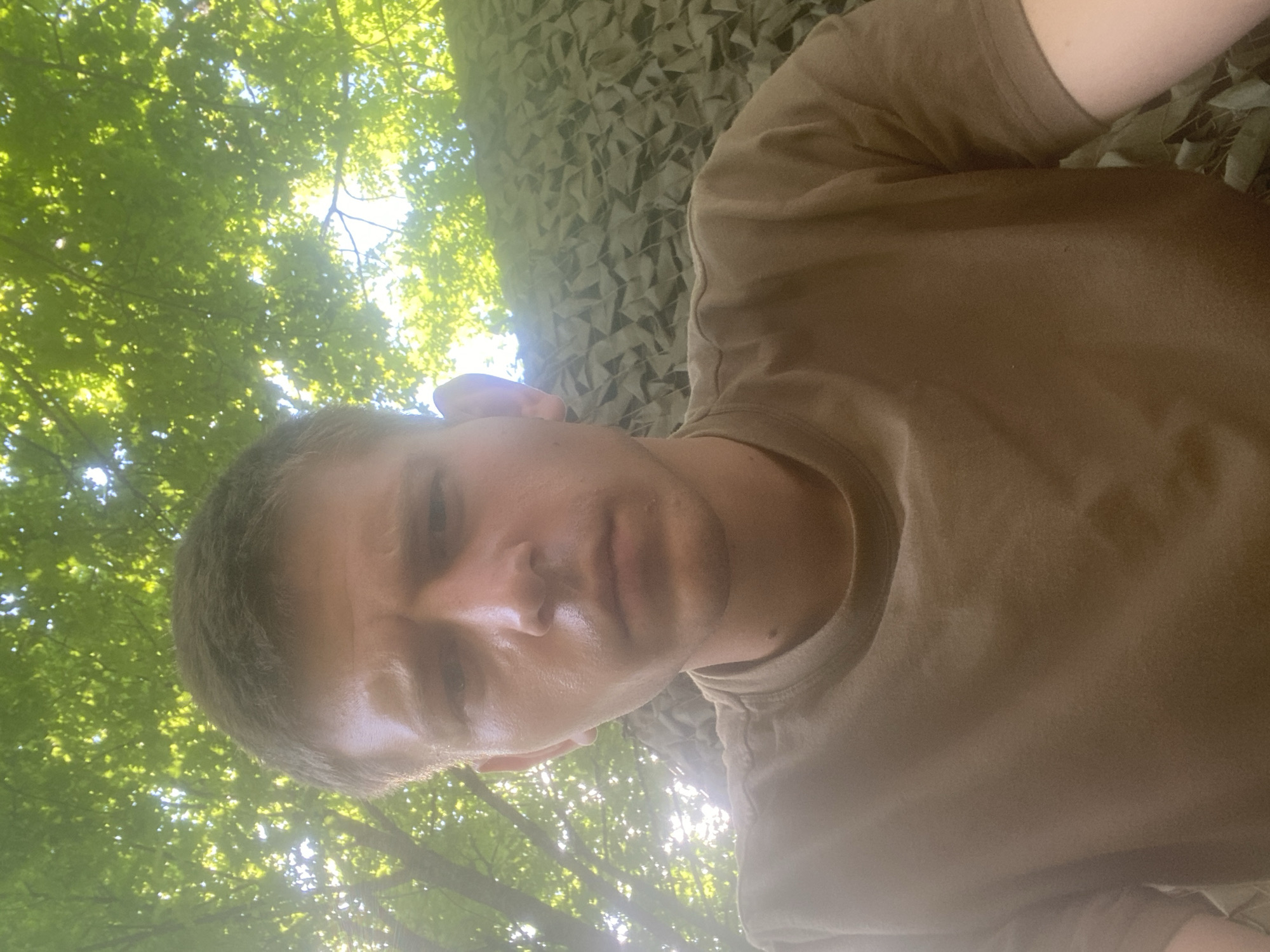 A young man with short brown hair and fair skin is pictured in a close-up, low-angle selfie taken outdoors. He is wearing a plain brown t-shirt. His expression is neutral, and his gaze is directed towards the camera.
The background features lush green tree leaves illuminated by sunlight from above. To the right of the man, partially visible, is a camouflage netting with a geometric pattern, suggesting an outdoor or possibly military setting. The environment appears natural, with the presence of trees and foliage. No specific landmarks or buildings are visible that would indicate a precise location within Shelkovo, Russia. The lighting suggests it is daytime, likely late morning or afternoon due to the dappled sunlight filtering through the leaves.