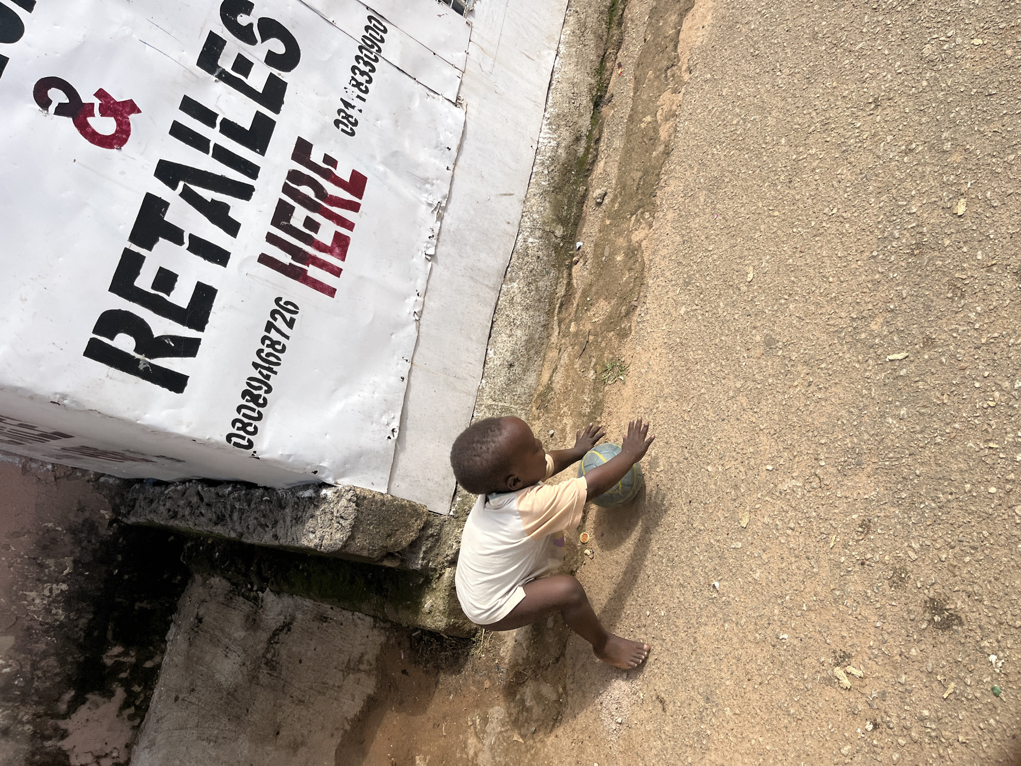 A young child, possibly a toddler, is crouched on a dusty, sandy ground in front of a white, corrugated metal structure. The child is wearing a light-colored t-shirt with peach-colored sleeves and no pants. The child is reaching for a small, multi-colored ball. The metal structure has black stencil lettering that reads "RETAILES" and "HERE" in red, along with phone numbers. The ground is uneven, with patches of dirt and some debris. To the left of the child, there is a concrete structure with visible cracks and some moss or algae growth. This scene appears to be in an outdoor, urban environment. The location is identified as the city of Jos, Nigeria, suggesting a context of a developing area within a city.
