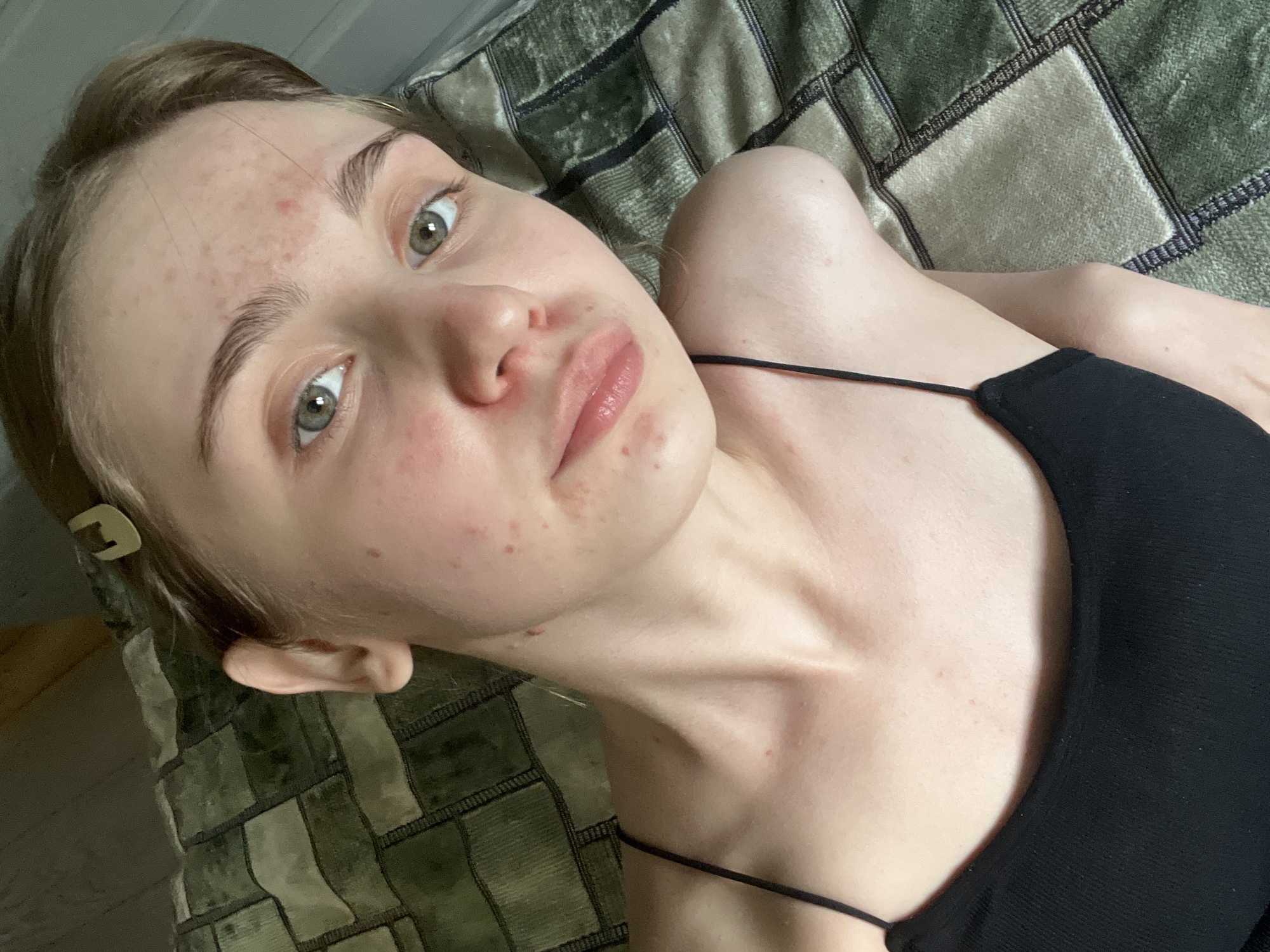 The image shows a close-up, overhead view of a young woman lying down, looking towards the camera. She has light brown hair pulled back with a clip, green eyes, and fair skin with visible acne. She is wearing a black spaghetti strap top. The background consists of a textured, dark green and light green plaid blanket. There are no indications of the city of Ashkadar, Russia, or any urban environment. The setting appears to be indoors.
