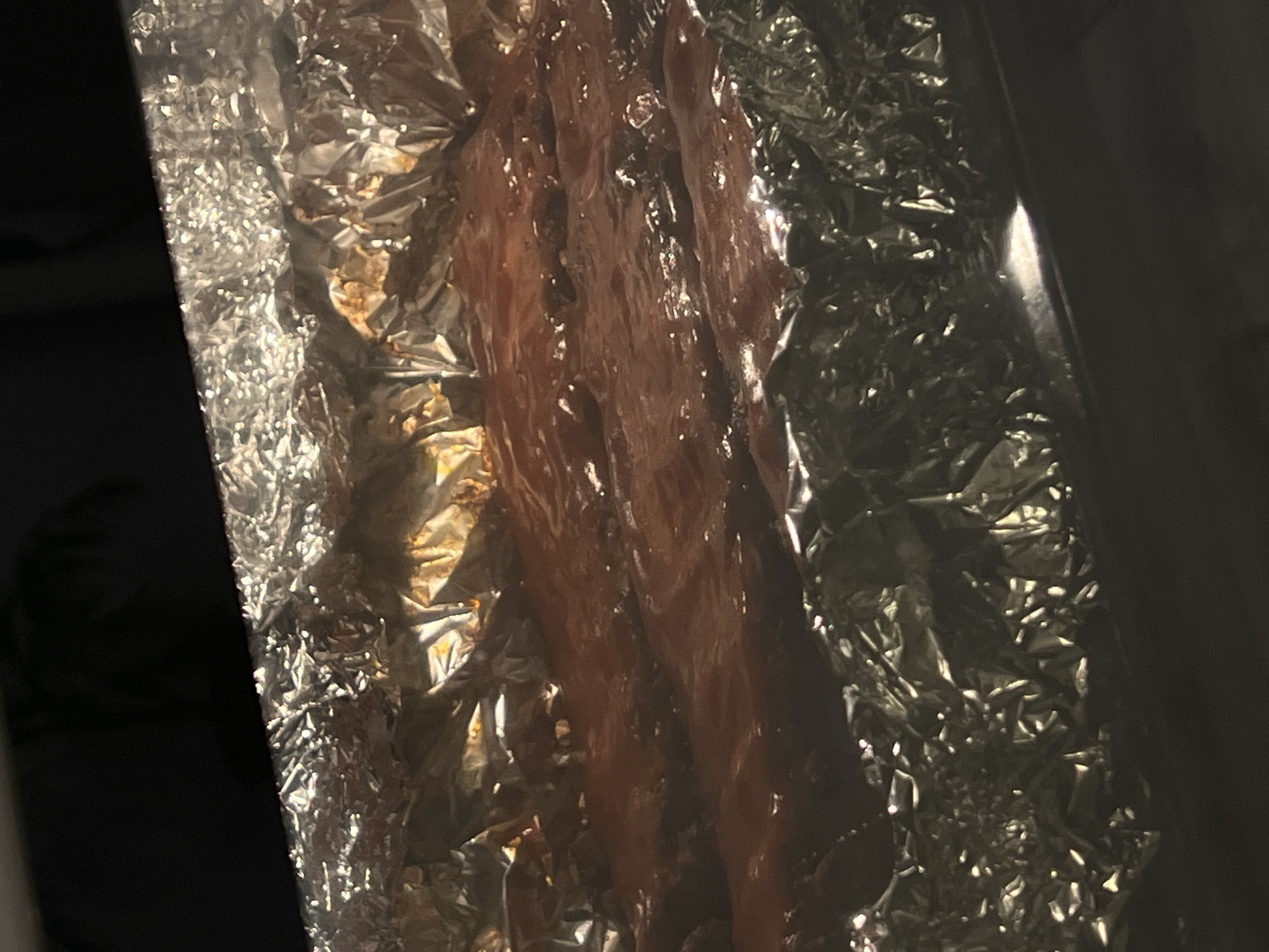 The image shows a close-up view of ribs covered in barbecue sauce, partially wrapped in aluminum foil. The ribs appear to be in a pan or baking dish. The lighting is dim, with some reflections on the foil. There are no people or other discernible objects in the image. The context of Florești, Romania is not depicted or implied by the visual information present.