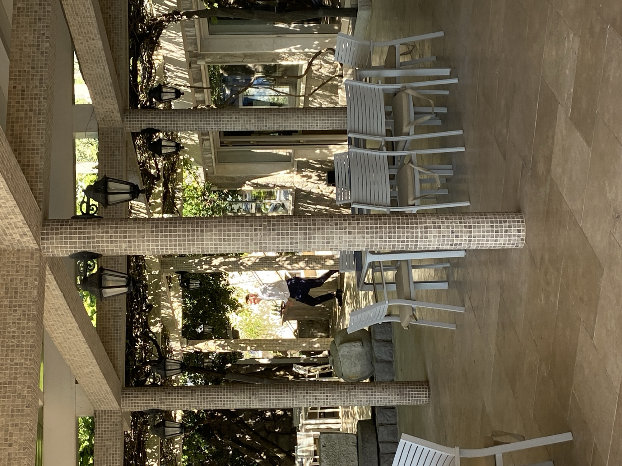 The image depicts a covered outdoor space, likely a patio or veranda, with multiple white chairs and tables. The structure is supported by columns covered in mosaic tiles, and hanging lanterns are visible. A person in a white shirt and dark pants is walking in the background. The location may be in Prijevor, Montenegro, but no specific identifying information is present in the image.