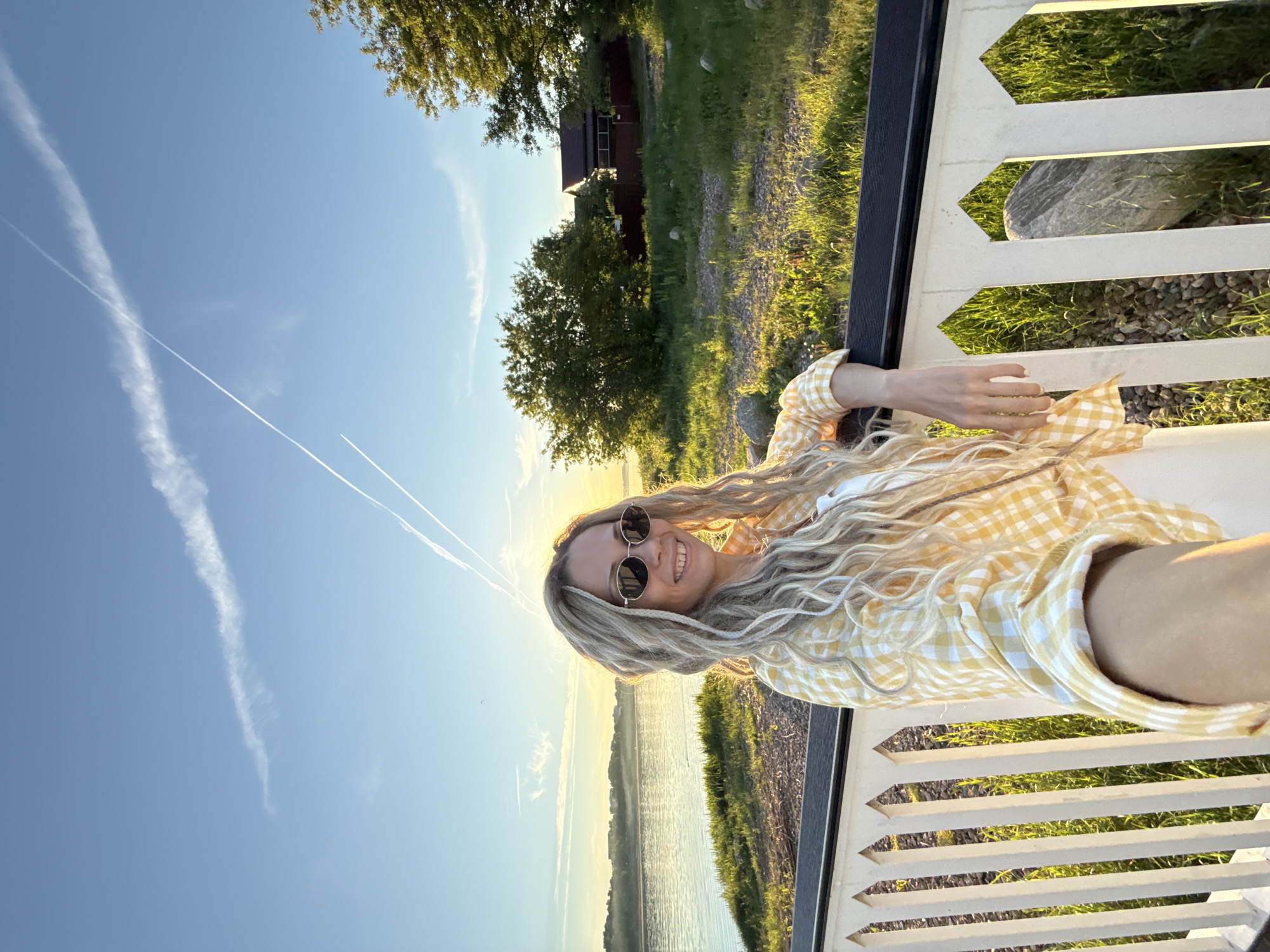 The image displays a waist-up, front-facing selfie of a female individual taken outdoors during golden hour or sunset, positioned along the bank of the Volga River in Yaroslavl, Russia.
The subject is a fair-skinned female with long, wavy, light-colored hair, styled in what appear to be dreadlocks or braids. She is smiling, revealing teeth, and wears oval-shaped dark sunglasses with thin metallic frames. Her attire consists of a yellow and white gingham (plaid) long-sleeved button-up shirt over a white undershirt, with white pants partially visible. Her left arm is extended towards the camera, holding a device, while her right forearm rests on the black top rail of a white decorative railing. Her fingernails display a light polish.
The white railing features vertical pickets with pointed arch-like designs at their tops. Behind the railing is a grassy area interspersed with scattered light-colored rocks and patches of bare earth, all illuminated by warm, low-angle sunlight.
Behind the subject and the railing, a wide river reflects the warm light from the low sun. The opposite riverbank presents a distant, hazy silhouette of what appears to be an urban or suburban area, including structures and vegetation. On the immediate bank to the subject's right, several mature deciduous trees are visible, along with a house or cottage featuring a reddish-brown roof and dark walls, partially obscured by foliage and a dark fence.
The sky is clear blue with two prominent white contrails from aircraft: one diagonally crosses the upper sky from top-left to mid-right, and another intersects it from upper-right towards mid-left. A small dark bird is visible in the sky above the distant horizon. The low sun, positioned out of frame to the upper right, casts a strong backlight, creating a warm glow around the subject and scene.